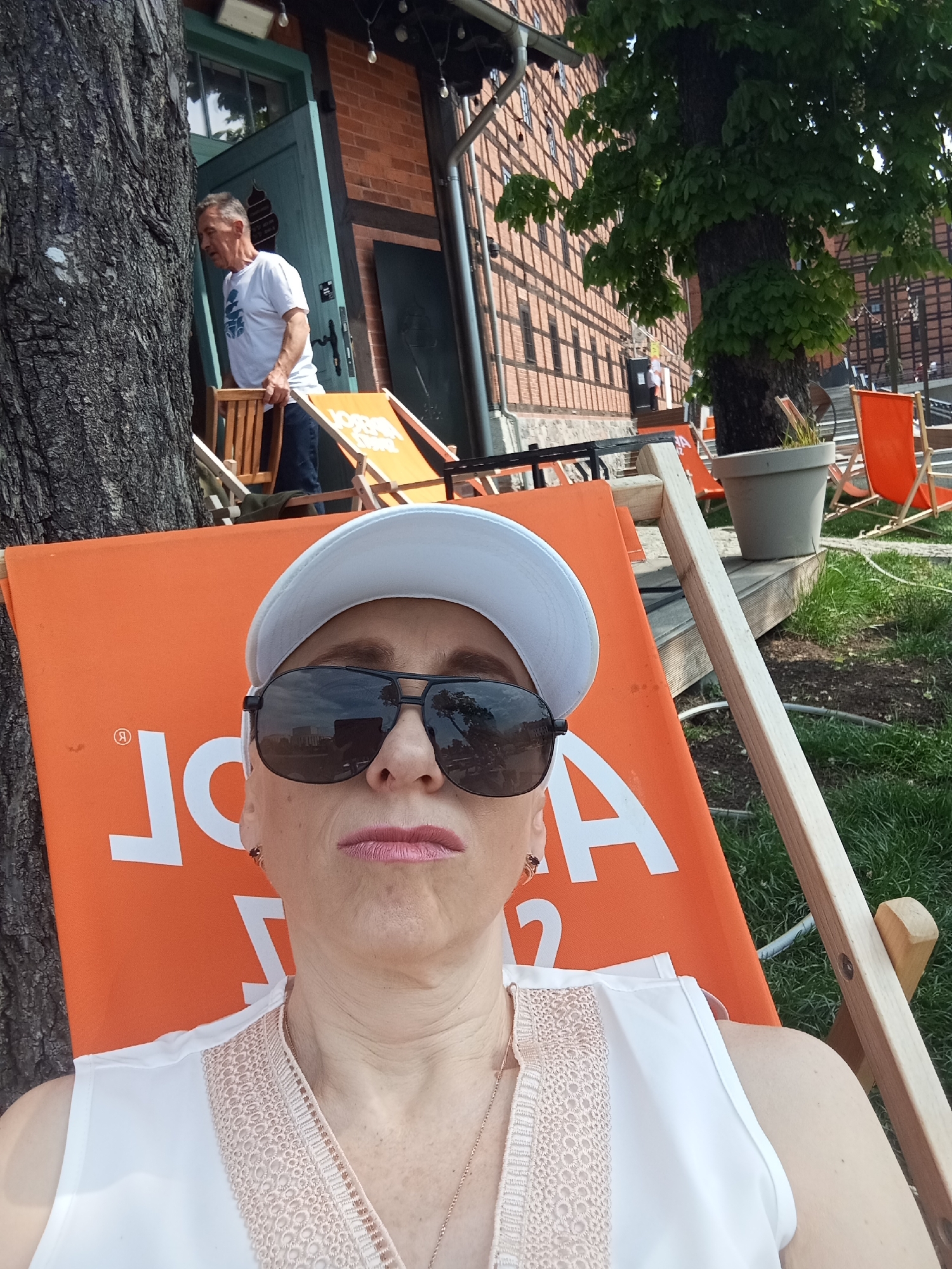 A woman wearing sunglasses and a white cap is seated in an orange beach chair. The chair is in a grassy area with other chairs visible in the background. A tree trunk is to the left of the woman, and a building with red brick and a teal door is in the background. A man in a white t-shirt and blue jeans is seen carrying a chair near the door of the building. The scene appears to be in the city of Bydgoszcz, Poland, given the architectural style and the presence of beach chairs, likely near a waterfront area.