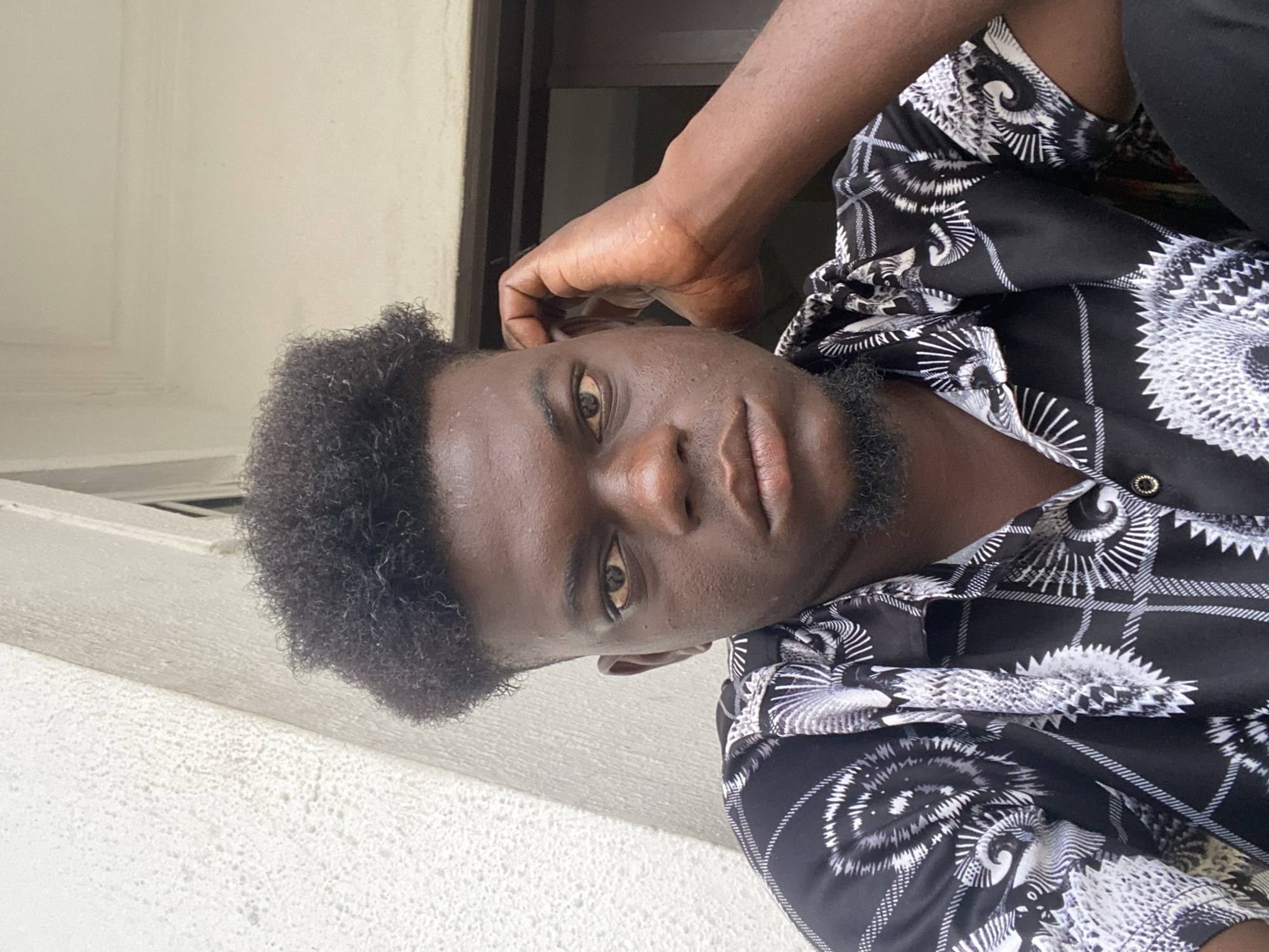 A close-up, portrait-oriented image shows a young Black man looking directly at the camera. The image is rotated 90 degrees counter-clockwise, making the man's head appear tilted to the right. He has dark, curly hair and a short, dark beard or stubble. His eyes are brown, and his facial expression is neutral to slightly serious. His skin has a noticeable sheen, possibly indicating sweat or oil.
He is wearing a black collared shirt with an intricate white and grey pattern that features abstract shapes resembling sunbursts, waves, or tribal motifs. A single metallic button, possibly gold or brass, is visible on his shirt. His right hand is raised to the side of his head, with his fingers near his right ear or temple, suggesting a casual or contemplative pose.
The background is out of focus, consisting of a light-colored, textured surface (possibly a wall or building exterior) on the lower and left side of the frame, and a darker, blurred rectangular shape (perhaps a window or doorway) on the upper and right side. The lighting suggests it is daytime. There is no visible text in the image.