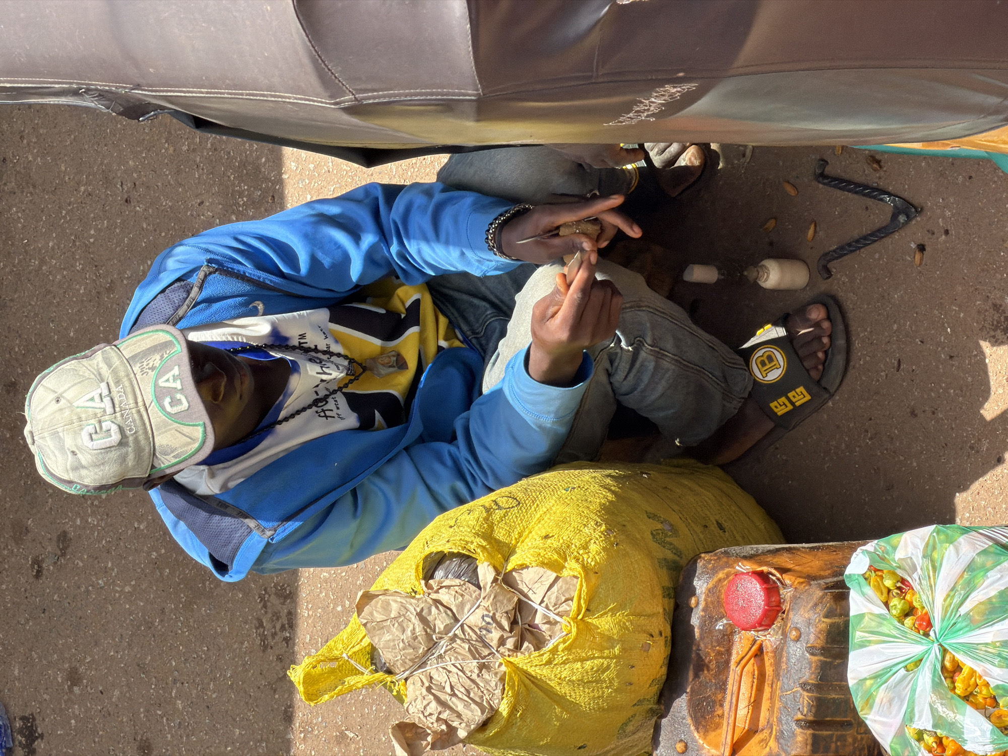 In Bukuru, Nigeria, a young person wearing a blue jacket, jeans, and a "CA CANADA" baseball cap is crouched on the ground next to a large, bright yellow bag. The person is holding a small object in their hands, appearing to be working with it. Scattered around them are various items, including a plastic container with a red cap, a bag of peppers, a small bottle, and a metal tool resembling a nutcracker. The ground is dusty and appears to be a street or market area.