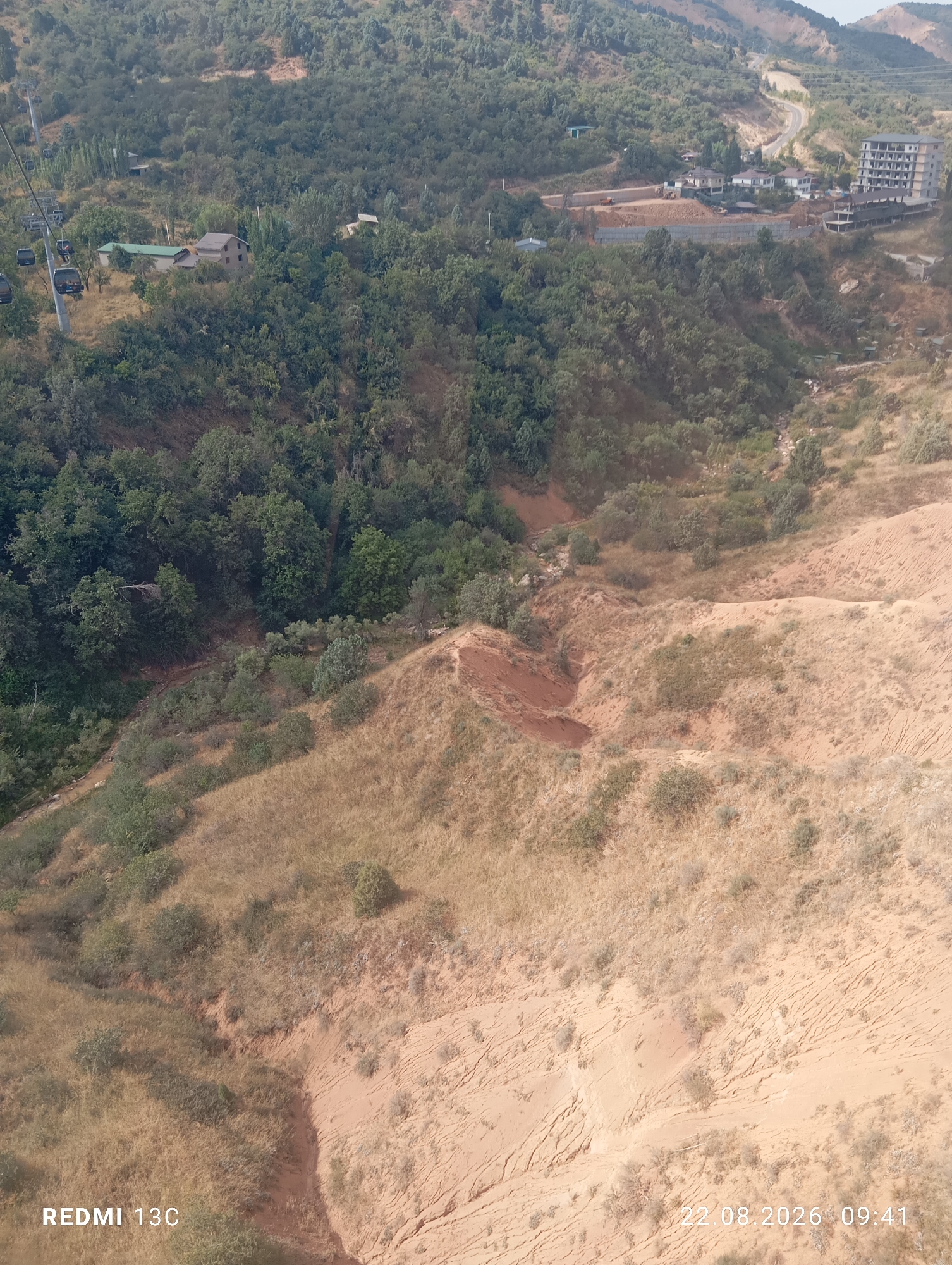 The aerial view depicts a mountainous landscape with a cable car system in the upper left corner, carrying several cabins. Below the cable car, a cluster of houses is nestled amongst dense green trees. Further into the scene, a winding road is visible, with buildings and construction sites dotting the hillside. The terrain transitions to dry, sandy slopes with sparse vegetation, characteristic of a semi-arid environment. The overall impression is of a resort or recreational area within a natural setting. The image is dated 22.08.2026, 09:41 and has "REDMI 13C" watermark.