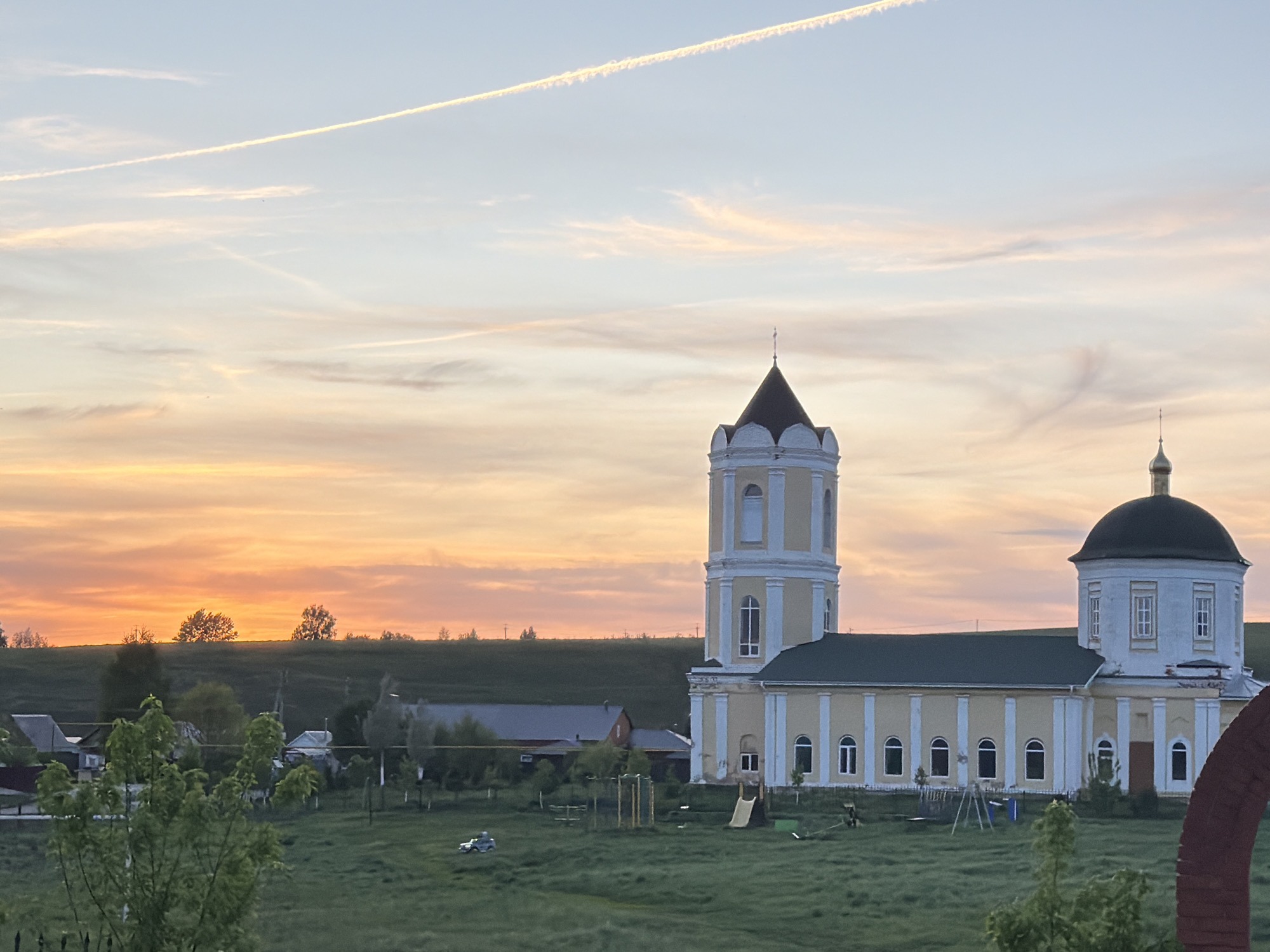 The image presents a rural scene likely captured near Ильбухтино, Russia. Dominating the view is a cream-colored church with a dark green dome and steeple. The church stands on a grassy field, which also contains a playground with a slide and swings. Behind the church are a few low buildings. A horizon of slightly elevated land is topped by trees. Above, the sky displays a sunset with hues of orange, pink, and pale blue, streaked with contrails.