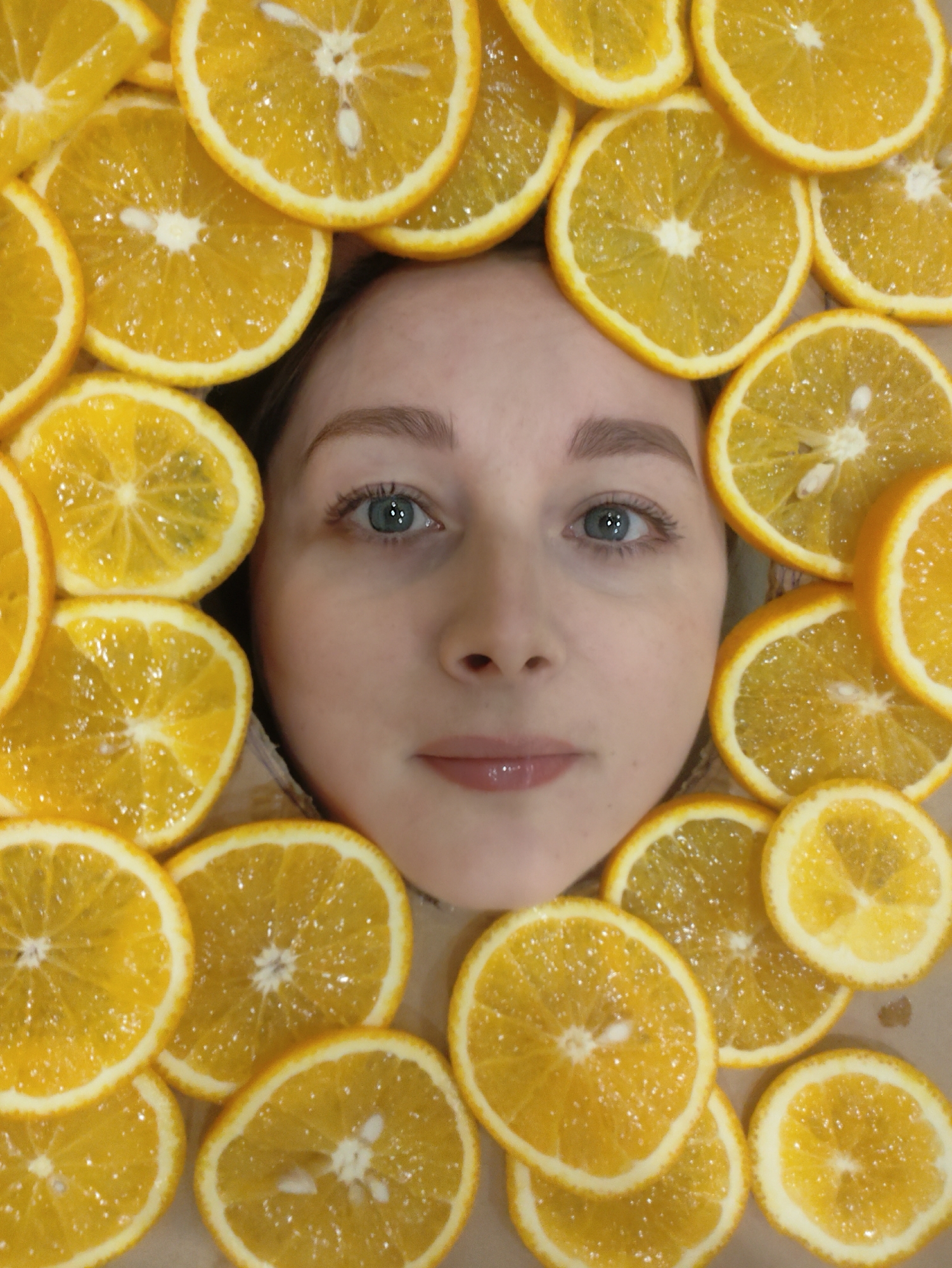 A person's face is framed by orange slices. The person has light skin, light brown hair, and blue eyes. The orange slices vary slightly in size and color, forming a border around the person's face. The setting is unknown.