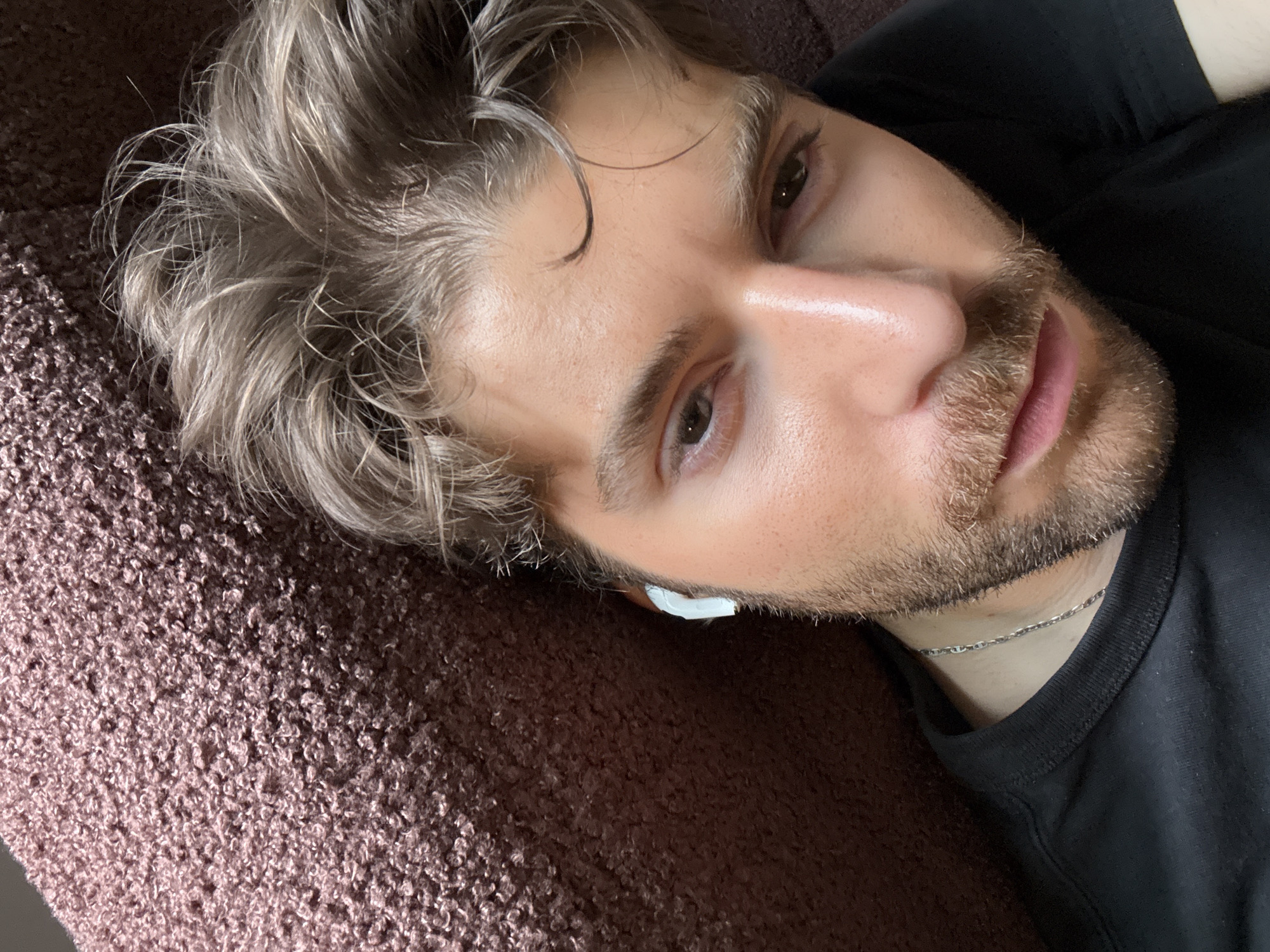 A close-up, slightly high-angle shot features a man lying down indoors, looking directly at the camera. He appears to be in his mid-20s to 30s, with fair skin, light brown or ashy blonde hair, and a light stubble beard. His brown eyes are open and meet the viewer's gaze. He is wearing a black t-shirt and a thin silver chain necklace. A white earbud is visible in his right ear. His head rests on a thick, textured fabric, possibly a blanket, couch, or bedspread, which is a mix of brown and purple hues. The lighting suggests daytime. His hair is casually styled, showing some volume and slight waves. His expression is neutral and calm. There is no visible text in the image. The setting appears intimate and informal.