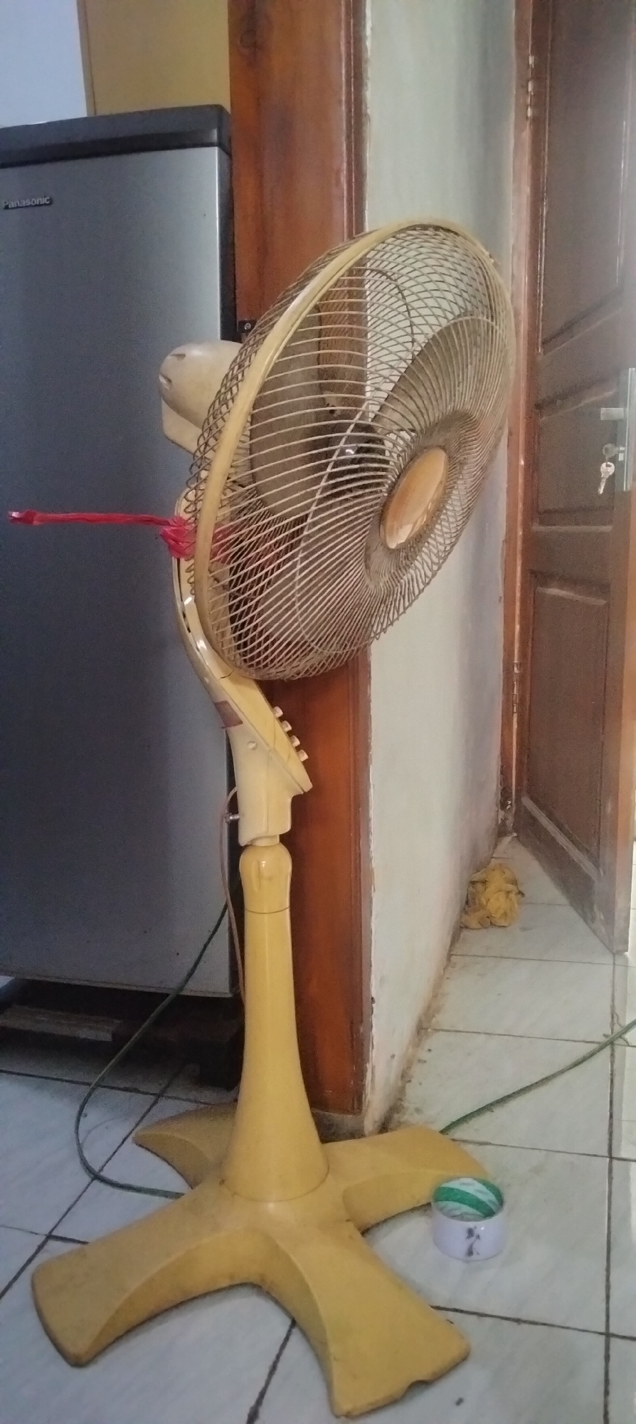 An upright yellow floor fan with a damaged oscillating mechanism, secured with red tape, stands on a tiled floor next to a grey Panasonic refrigerator. The fan has a wire mesh grill and a circular control panel with buttons. A wooden door frame is visible behind the fan, with a yellow mop ball in the corner. The scene is indoors, likely in a residential setting in Antirogo, Indonesia, given the context of the location.