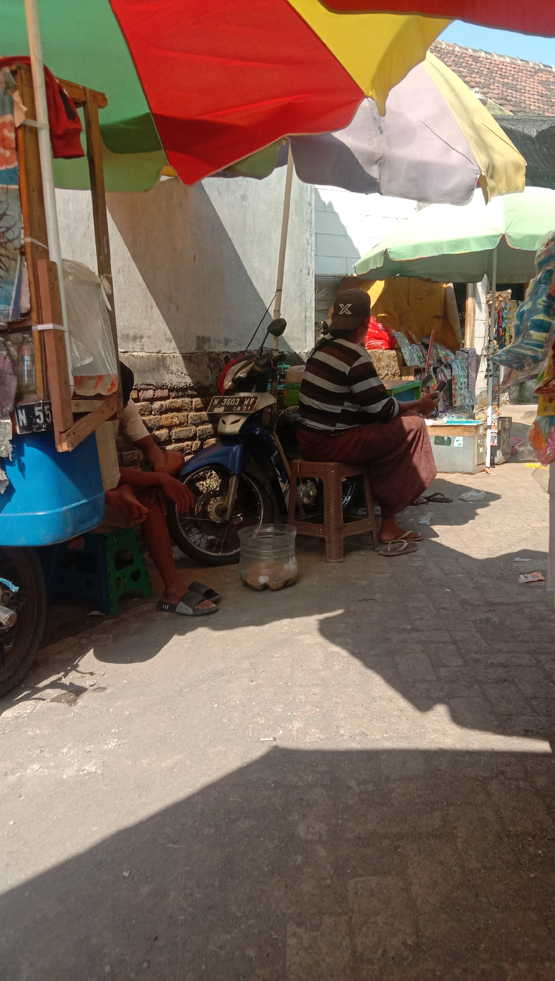 A sunny outdoor scene in Karanganyar, Indonesia shows two men under colorful umbrellas at a roadside stall. The man on the left is seated on a small green stool next to a blue and white cart. He wears a light-colored shirt and dark trousers. The man on the right is seated on a brown plastic stool, facing away from the camera, wearing a striped shirt and dark sarong. He holds a mobile phone in his right hand. A blue motorcycle is parked behind the man on the left. In front of the motorcycle and between the two men is a clear plastic container holding what appears to be food items. The ground is paved and casts long shadows from the umbrellas and objects. In the background, a brick wall and buildings with tiled roofs are visible. The scene depicts a typical roadside vendor setup in an Indonesian town.