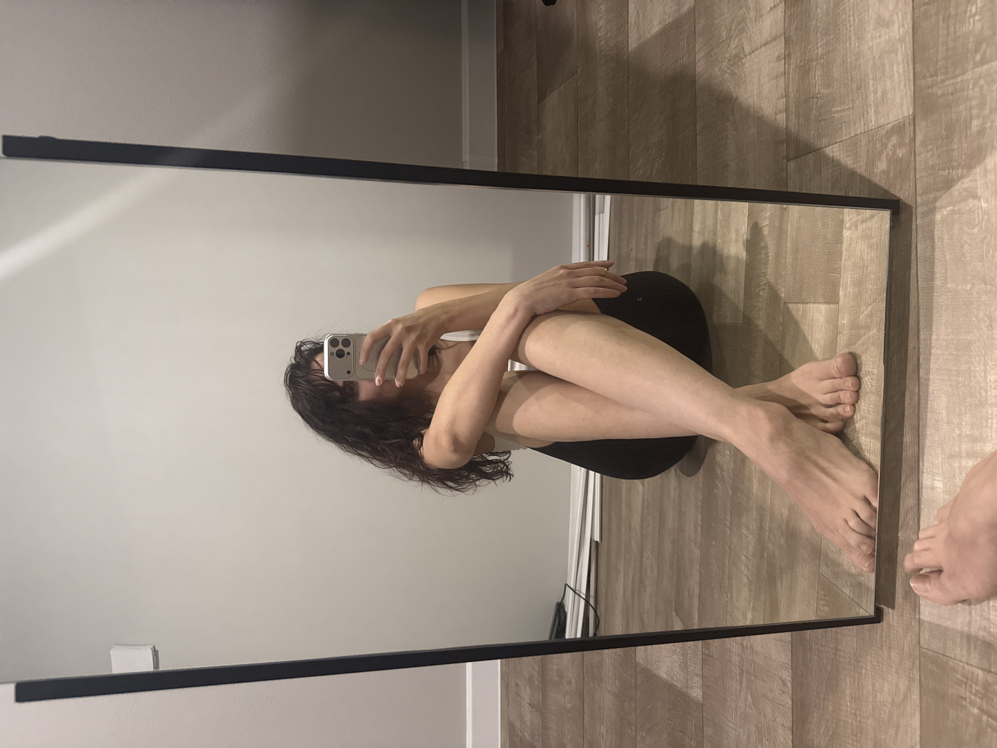 A reflection in a rectangular mirror shows a person sitting on the floor with knees bent, arms wrapped around legs, and head down, obscured by dark, curly hair and a cell phone being held up. The person is wearing what appears to be a light-colored top and dark bottoms. The background of the reflection shows a light gray wall. Outside the mirror, the floor is visible and appears to be wood-grain laminate. A single bare foot is partially visible at the bottom right corner of the frame. The location is unknown.