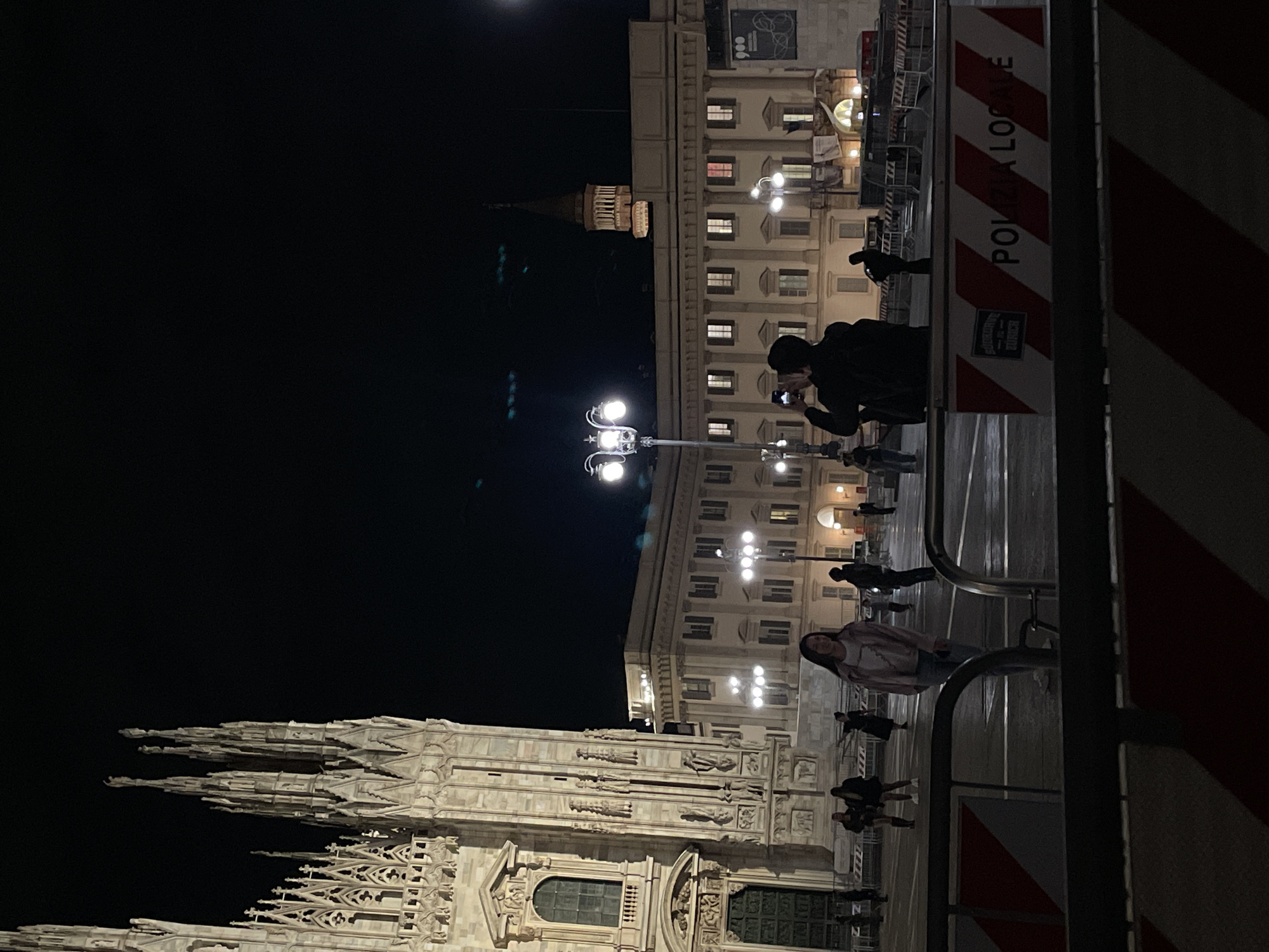 The image shows a nighttime scene in Milan, Italy, featuring the Duomo di Milano in the lower left. The cathedral's spires and facade are partially visible. Several people are present, some walking and others taking photos. A person is seen holding a camera and pointing it towards the buildings. In the background, another building with illuminated windows can be seen, partially obscured by streetlights. A "Polizia Locale" (Local Police) barrier is visible in the foreground on the right side of the image.