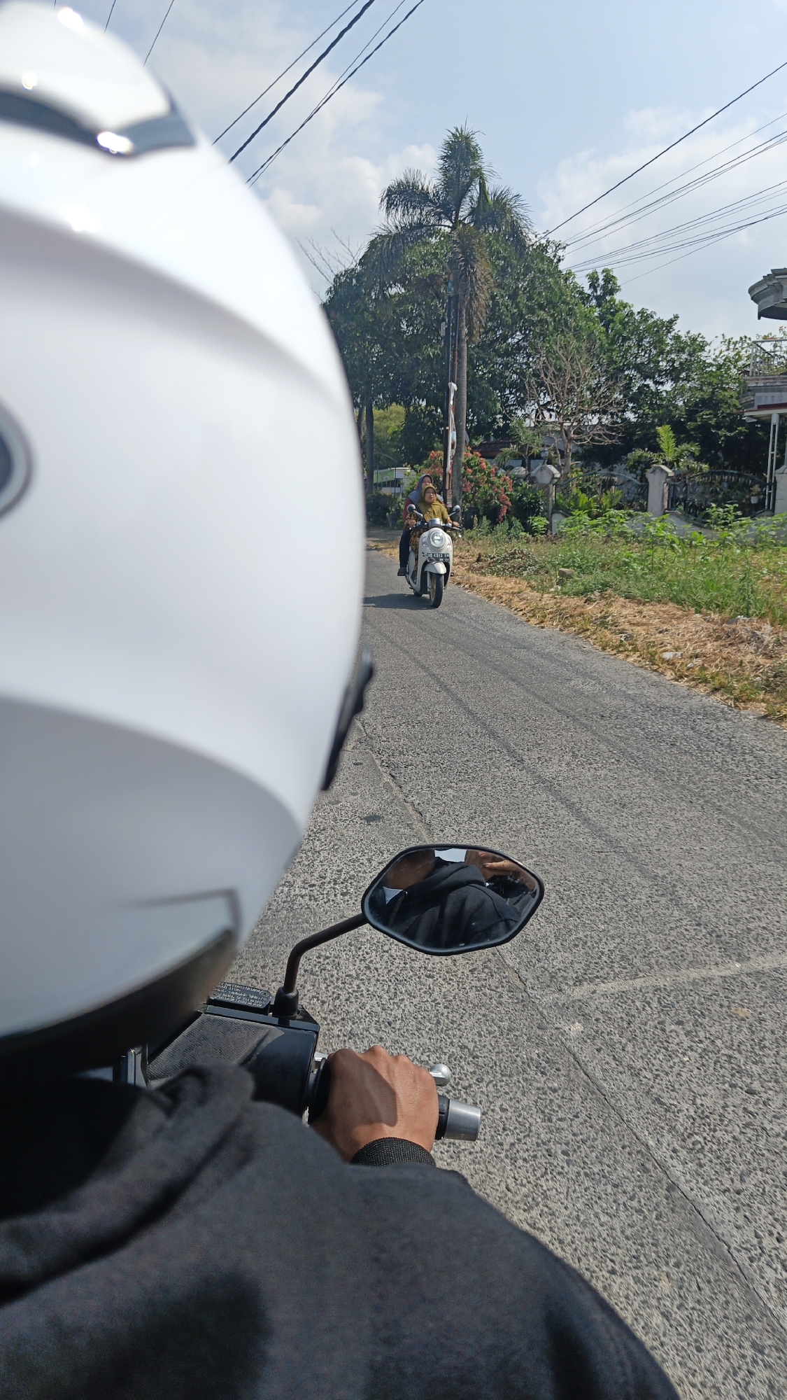 The image depicts a first-person perspective of someone riding a motorcycle on a paved road in Desa Sobokerto, Indonesia. The rider is wearing a white helmet and a black jacket, with their right hand visible on the handlebar. A rearview mirror reflects the rider's black jacket. Ahead on the road, another motorcycle is visible carrying two individuals, a driver and a passenger, both wearing light-colored clothing and headscarves. The road is lined with vegetation and trees, including a prominent palm tree. Power lines are strung across the clear blue sky. The environment appears to be a rural or suburban area with modest buildings visible in the background. The lighting suggests it is daytime, likely sunny.