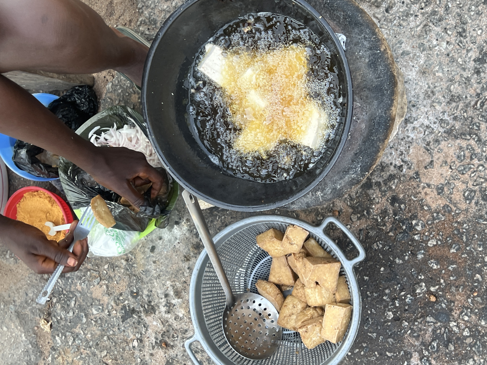The image depicts an outdoor food preparation scene in Jalingo, Nigeria. A dark-skinned individual, whose hands and lower right leg are visible, is engaged in deep-frying food. The person's right hand holds a small metallic fork and tongs while interacting with food inside a clear plastic bag, which is partially encased in a black plastic bag.
The central focus is a large, dark, round pot sitting on a low, dark stand, containing actively bubbling hot oil. Several white, cuboid pieces of food, likely tofu or bean cakes, are submerged in the oil, rapidly frying and developing a golden-brown crust.
To the left of the frying pot, a grey plastic colander holds a mound of previously fried, golden-brown, irregularly shaped cuboid pieces of the same food. A metallic slotted spoon rests within the colander.
In the upper background, several items are visible: a red bowl contains a heap of fine, orange-yellow powder, along with a small white plastic spoon. Next to it, a blue bowl holds a crumpled black plastic bag. Partially visible is another container holding thinly sliced white onions.
The individual's lower right leg, clad in a dark sandal, is visible near the frying setup. The entire operation is conducted on a rough, dark grey ground surface, consistent with concrete or asphalt, showing varying textures and small aggregates.