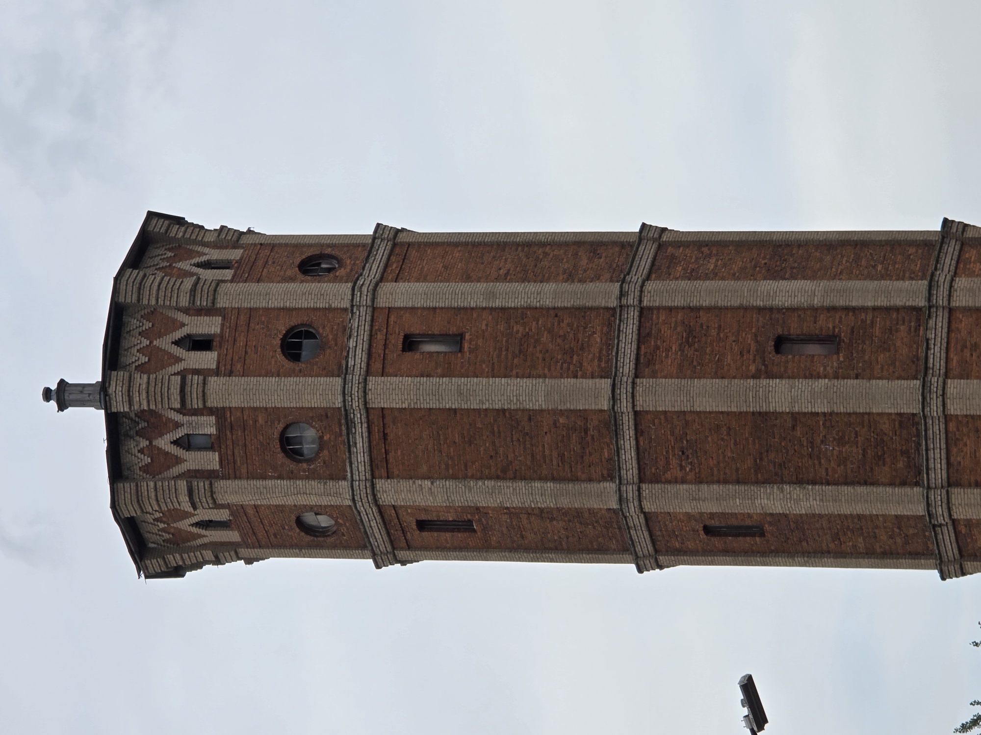 The image shows a close-up, low-angle view of a tall, octagonal brick water tower against a cloudy sky. The tower is constructed from dark red bricks with lighter colored brick accents forming vertical bands and decorative elements near the top. Several windows are visible: small, arched windows near the top, circular windows on the next level down, and narrow, rectangular windows on the lower sections. The top of the tower features a decorative cornice and a small metallic structure. To the left of the tower, a street lamp is visible. The scene is devoid of people and appears to be an exterior shot of an architectural structure in an urban setting. It is not possible to determine the specific city, let alone Istanbul, Turkey, from the visual information provided.