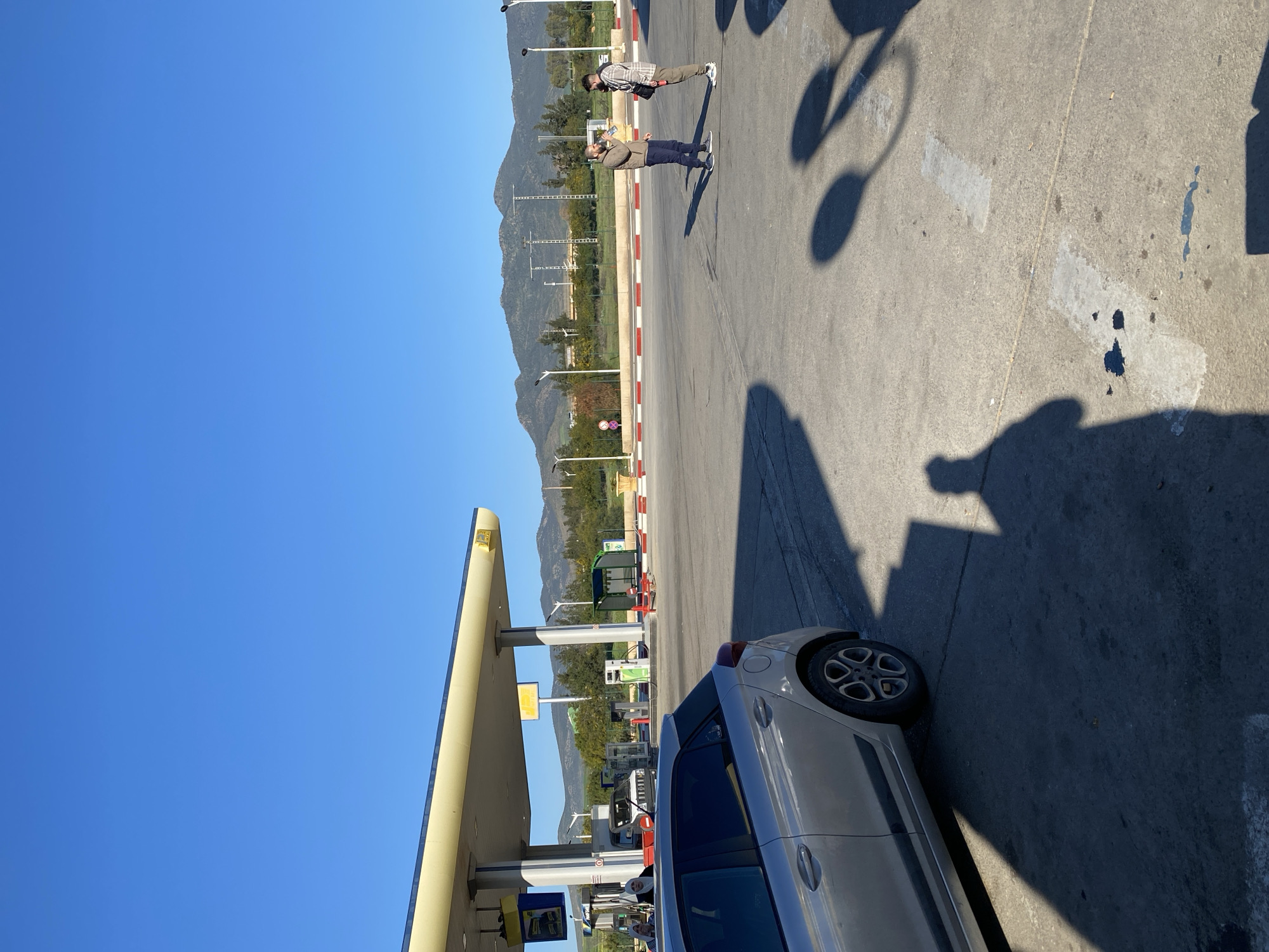 The image shows a scene at a gas station in Djelida, Algeria. A silver car is parked near the pumps. Two men are standing further away, one holding a phone up as if taking a picture. Mountains are visible in the background, beneath a clear, blue sky. Shadows are cast across the paved ground, including the shadow of a light fixture. Red and white stripes mark the edge of the parking area.