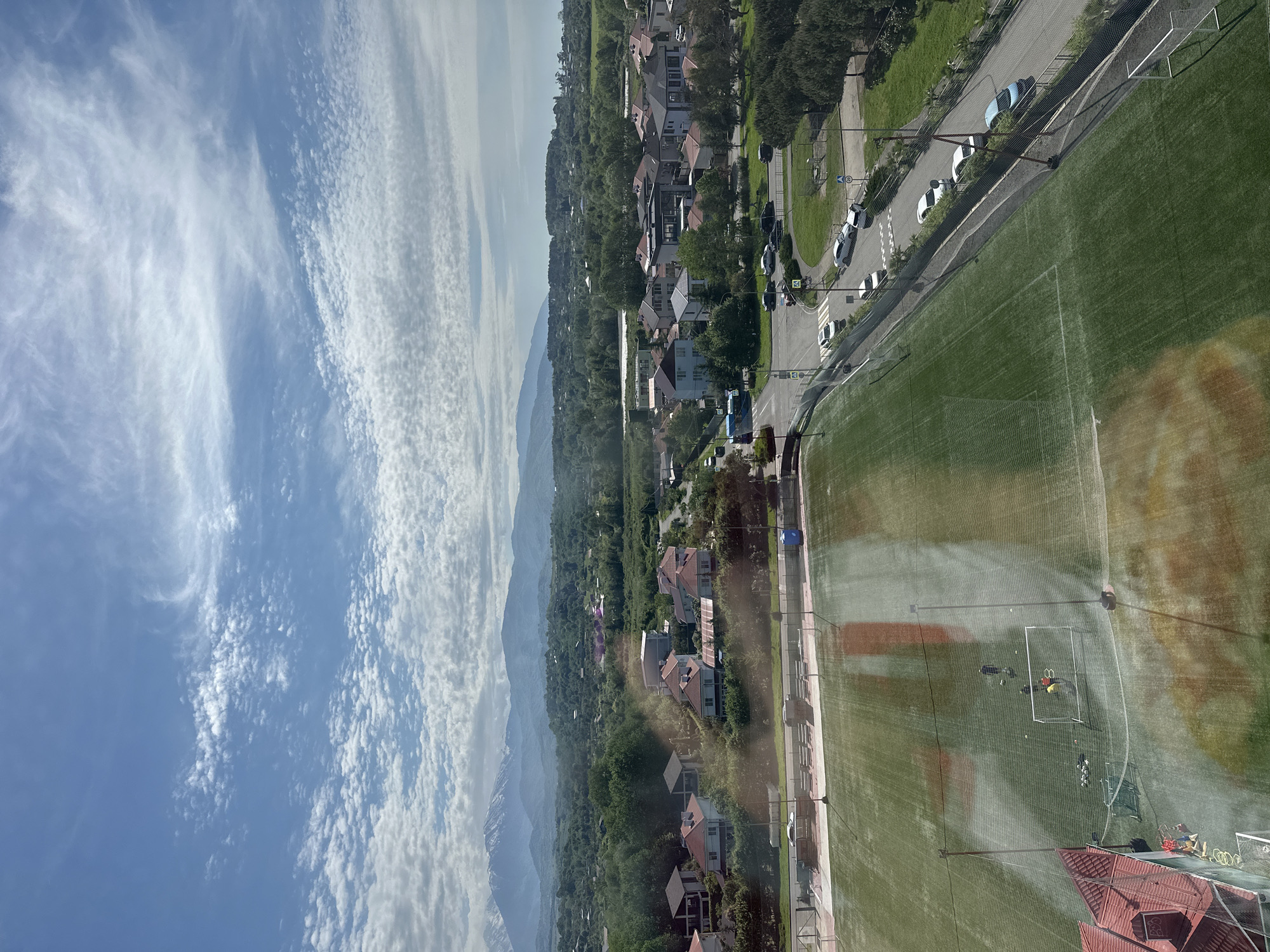 The image shows a soccer field adjacent to a residential area and a road, with mountains in the background and a cloudy sky above. People are visible on the soccer field near one of the goals. Cars are parked along the road next to the field. Houses are situated among trees. The image seems to be taken from an elevated viewpoint, possibly a building. It is not possible to determine the location of the image as Sochi, Russia from the visible context.