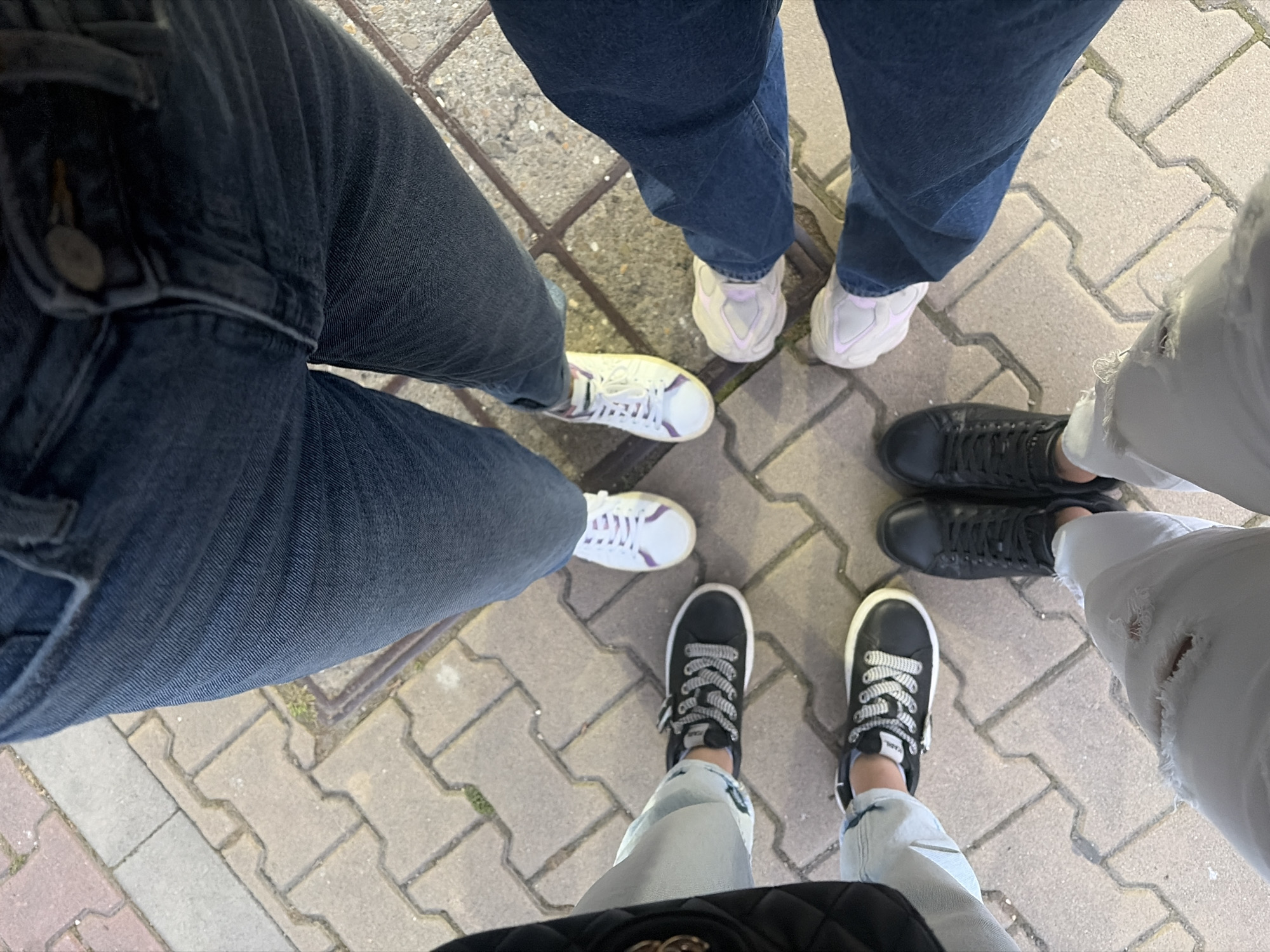 A low-angle, medium shot captures the lower halves of at least six people standing in a circle on a paved walkway composed of rectangular grey and tan interlocking bricks. The people are wearing jeans and sneakers, and the focus is on their footwear and legs.
Starting from the top and moving clockwise, the first person is wearing dark blue jeans and white sneakers with purple accents. The second person is wearing dark blue jeans and light pink chunky sneakers. The third person is also wearing dark blue jeans and light pink chunky sneakers. The fourth person is wearing light gray, ripped jeans and black sneakers. The fifth person is wearing light blue jeans and black sneakers with white laces. The sixth person is wearing light blue jeans and black sneakers with white laces.
The walkway appears to be outdoors, possibly in a city. The lighting suggests it is daytime, with some shadows present. The background is out of focus, but the brick pattern of the walkway is clear. No faces or upper bodies of the individuals are visible.
