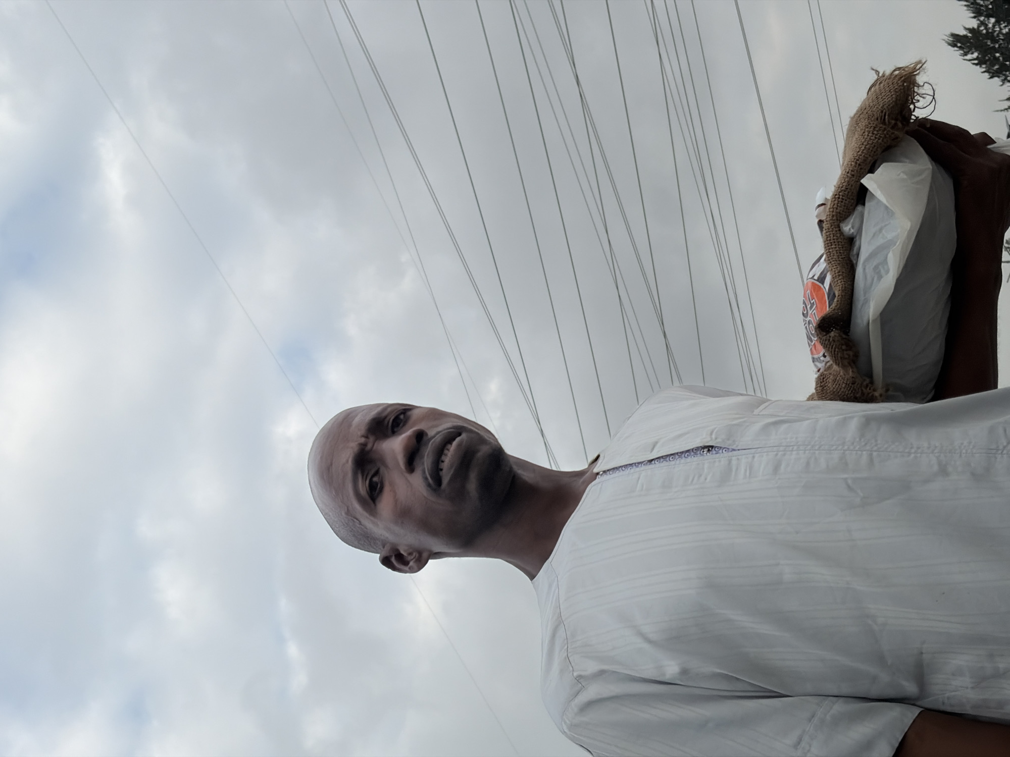 A medium shot captures a bald Black man in Igboefon, Nigeria, wearing a white traditional tunic with pinstripe texture. He is looking directly at the camera with a determined expression, his lips slightly parted. The man is holding a bundle wrapped in white plastic and burlap. The background is dominated by a cloudy sky and a network of power lines stretching across the frame. The lighting is soft, suggesting an overcast day.