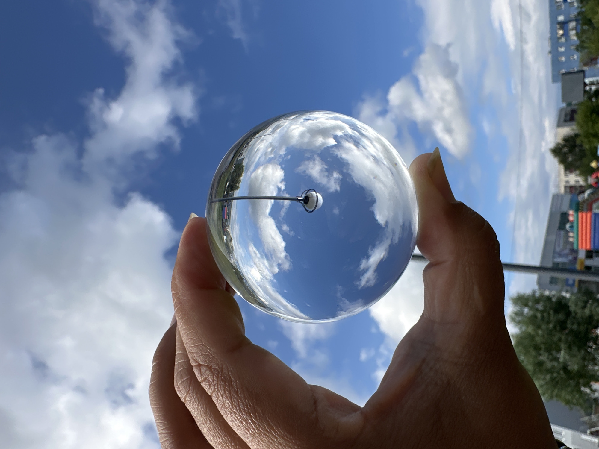 This media file features a close-up shot of a hand holding a clear glass sphere, which acts as a lens, inverting and reflecting the surrounding environment. The sphere captures an upside-down view of a bright blue sky filled with fluffy white clouds. A thin metal rod with a small, spherical attachment extends from the center of the sphere, also inverted.
The background, seen through and around the sphere, depicts a sunny day with a clear blue sky and scattered clouds. To the right of the sphere, the scene is inverted, showing buildings, trees, and what appears to be a colorful, possibly recreational area with some signage. The time of day is clearly daytime, indicated by the bright sunlight. The weather appears to be pleasant and fair, with a mix of sun and clouds.
The hand holding the sphere is in focus in the foreground, with visible skin texture and a thumb and forefinger gently cradling the glass. The overall mood conveyed is one of curiosity and perhaps wonder at the distorted perspective offered by the sphere. No visible text is present in the media file.