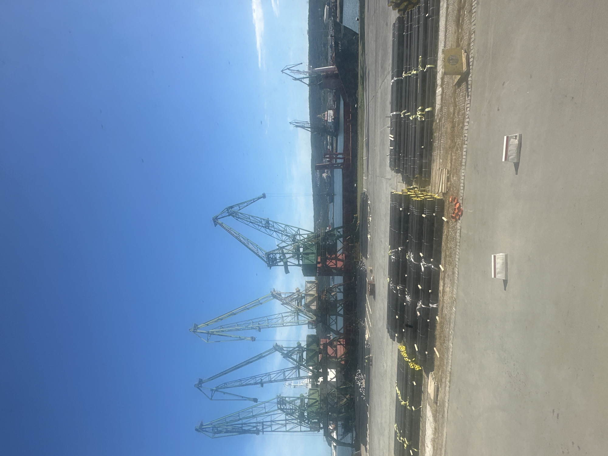 This is a medium shot, eye-level, outdoor, daytime photograph of a port facility. The sky is clear and blue. In the foreground, stacks of black cylindrical pipes are neatly arranged on a paved surface. Beyond the pipes, a harbor scene unfolds with large loading cranes, ships, and more industrial equipment. Several massive, green and red port cranes dominate the middle ground, positioned along a waterfront. One ship, labeled "YEBA," is docked and being serviced by a crane. In the background, rolling hills covered in vegetation are visible, suggesting a coastal or riverside location. The overall atmosphere is industrial and functional.