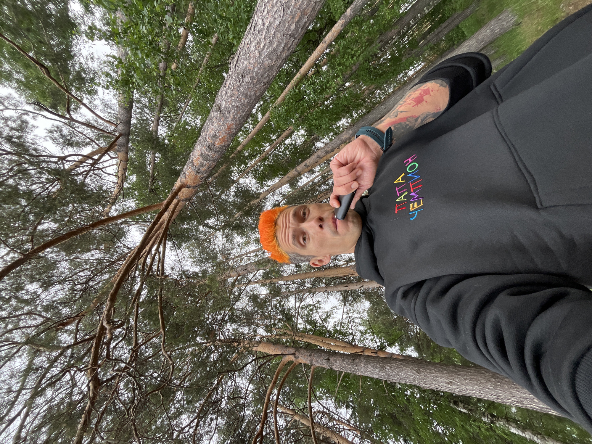 A male individual with bright orange, spiky hair and prominent stubble is depicted from a low-angle perspective in a wooded area within the city of Chaikovsky, Russia. He wears a black pullover hoodie featuring the embroidered Cyrillic text "ПАППА ЧЕМПИОН" (PAPPA CHEMPION) in multicolored letters across the chest. His left forearm displays a red and black tattoo, and a dark blue or green smartwatch is worn on his left wrist. The subject holds a black, cylindrical electronic vaping device to his mouth, appearing to be in the act of inhaling or exhaling. He looks directly upwards towards the camera with a neutral expression. The background consists of numerous tall pine trees with reddish-brown bark and green needles, along with some deciduous trees featuring green foliage, all reaching upwards towards an overcast sky. The overall lighting is diffused and natural.