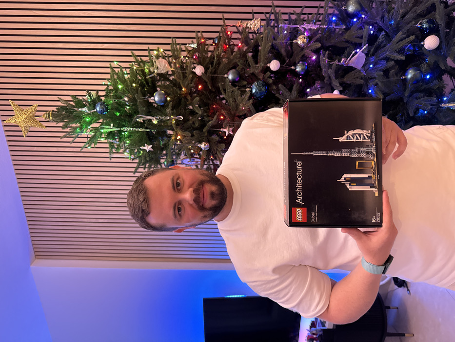 A light-skinned adult male, with short dark hair and a beard, wearing a white t-shirt and a light blue smartwatch, stands indoors, looking directly at the camera with a slight smile. He holds a black rectangular LEGO Architecture box, model 21052, labeled "Dubai," which displays miniature representations of the Burj Khalifa, Jumeirah Emirates Towers, and Burj Al Arab. These iconic structures are situated in Dubai, United Arab Emirates. To the man's right, an artificial Christmas tree is decorated with a gold star topper, spherical blue and white ornaments, icicle decorations, and multi-colored string lights. To his left, a dark television screen rests on a cabinet, with a blue ambient light emanating from behind it. The background features a light-colored wall with vertical wooden slat paneling.