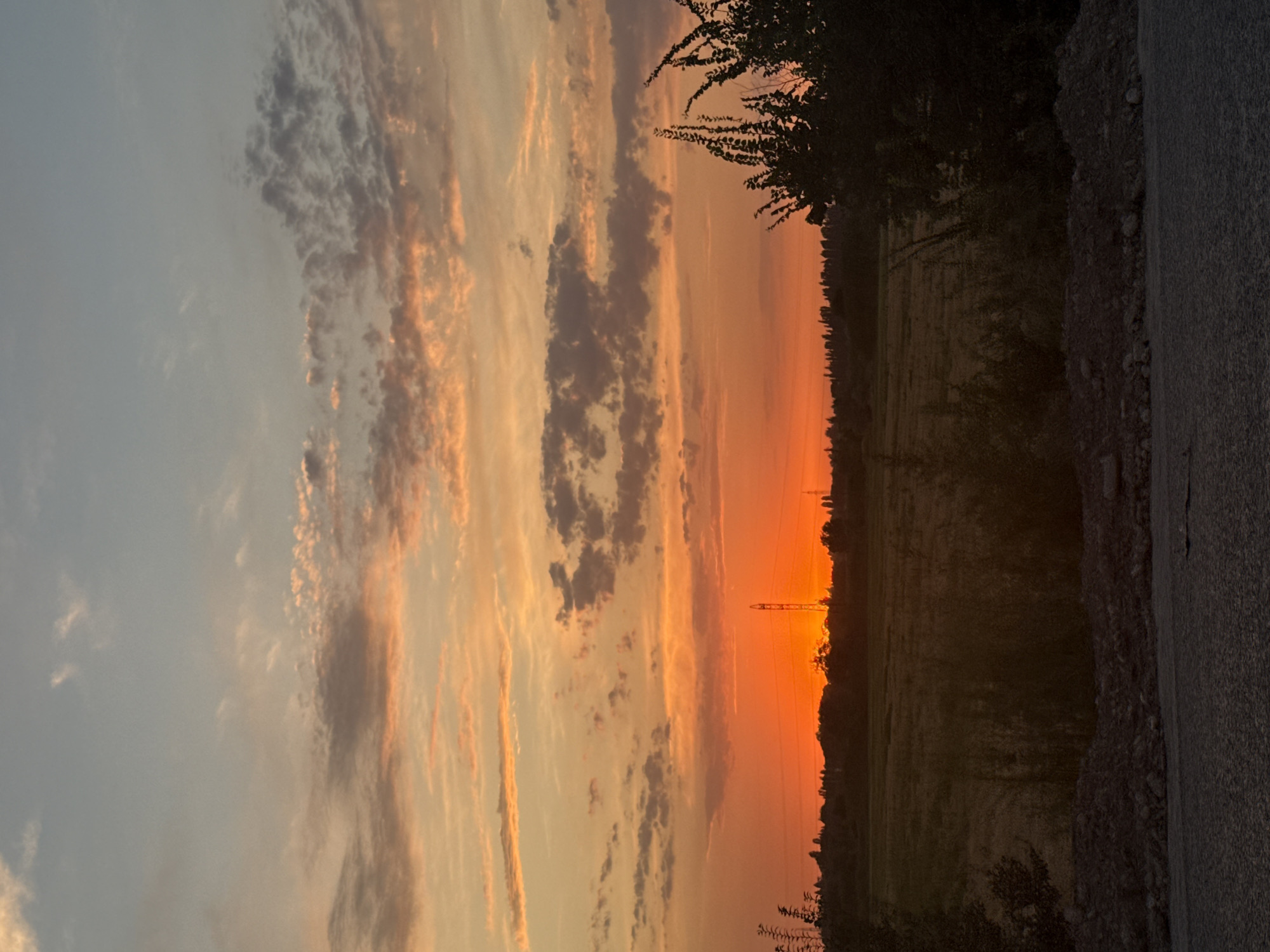 A vertical, eye-level shot captures a vibrant sunset over a rural landscape. The sky is a canvas of fiery orange and soft pastel hues, with scattered clouds tinged with pink and gray. A prominent orange glow emanates from the horizon, where the sun is setting behind a distant tree line. A tall, slender transmission tower stands silhouetted against the bright sky, its structure clearly defined. In the foreground, dark silhouettes of trees and bushes frame the scene, with a patch of textured ground, possibly dirt or gravel, at the bottom. The overall impression is one of serene natural beauty in a tranquil, undeveloped area, possibly on the outskirts of Drujba, Kyrgyzstan.
