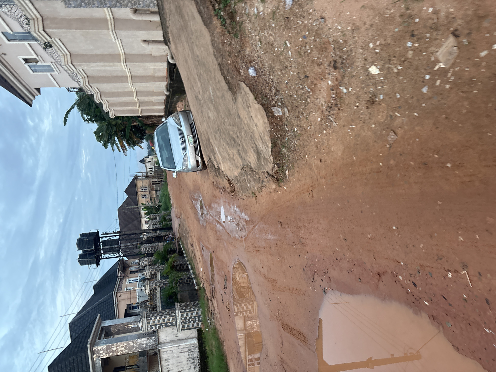 The image depicts a red, muddy road in Obosi, Nigeria, dotted with large puddles of water. A silver sedan is parked on the right side of the road, facing away from the viewer. The car appears to be on a slightly elevated concrete surface. On the left side of the road, a wall with decorative pillars separates the road from a residential area. Beyond the wall, several houses are visible, some with dark, pitched roofs and others with lighter facades. In the background, a metal structure supporting multiple large black water tanks stands tall. The sky is overcast with clouds. The overall scene suggests an environment characterized by unpaved roads and significant rainfall.