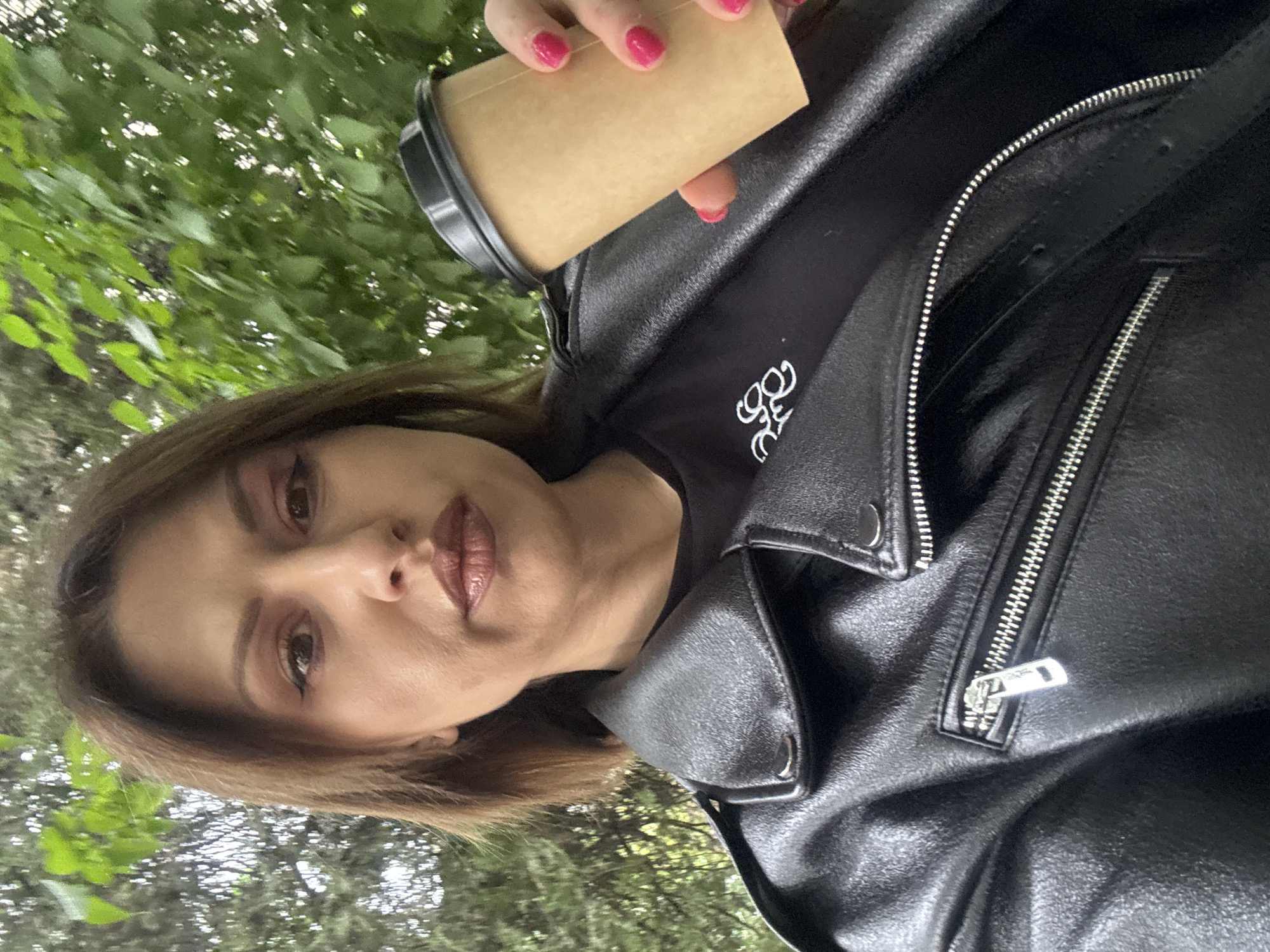A woman is holding a coffee cup, wearing a black leather jacket over a black shirt. The background is blurry and appears to be foliage, suggesting an outdoor location. The picture's angle is slightly tilted. The city of Sofia, Bulgaria cannot be determined from the image.