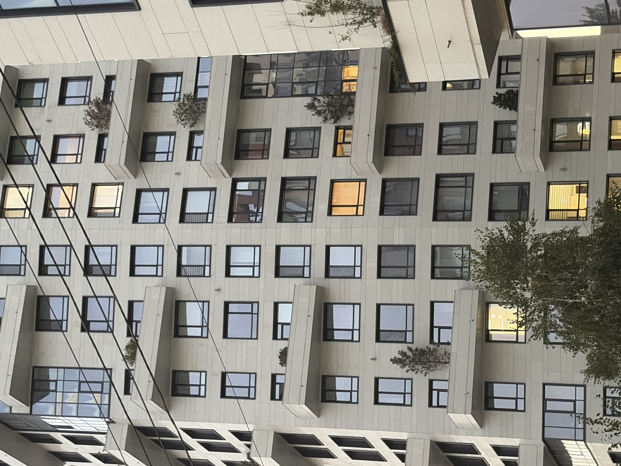 The image shows a modern, light-colored apartment building with numerous windows and balconies. Several windows are illuminated, indicating occupancy. Trees and greenery are visible on some balconies and at the base of the building. Power lines cross diagonally in front of the building. The architectural style suggests a contemporary urban environment. This building is located in Almaty, Kazakhstan.