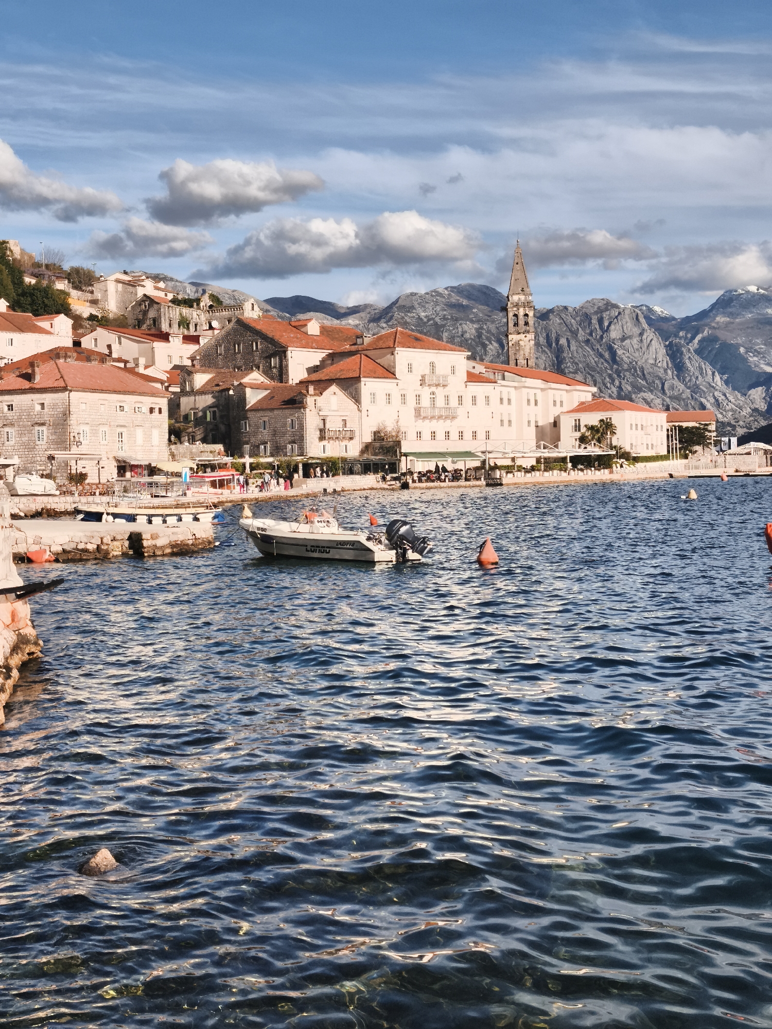 A daytime shot shows Perast, Montenegro. A townscape features buildings with red-tiled roofs and a tall church tower. The buildings sit along the shoreline. Mountains rise in the background under a sky with scattered clouds. In the foreground, water ripples with a boat floating in the middle and orange buoys dotting the surface.