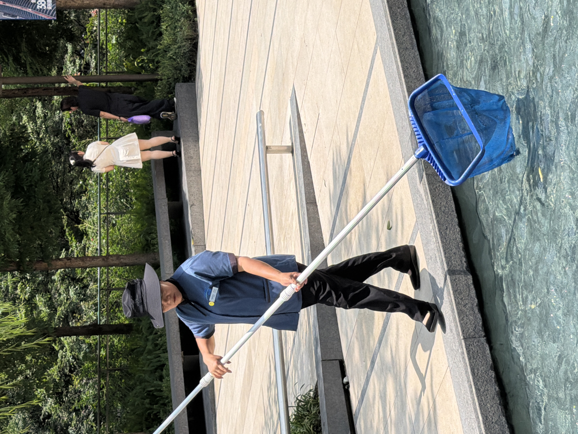 In Yuzhong District, China, a person wearing a blue uniform and a black wide-brimmed hat stands on a paved pathway next to a pool of water. They are holding a long pole with a blue net attached, which is partially submerged in the water. The person is looking down at the net, appearing to be in the process of cleaning the pool. In the background, two other individuals, one in a white dress and another in black clothing, are visible. The environment is outdoors, with trees and foliage providing a green backdrop. The paved area features sunlight casting shadows.