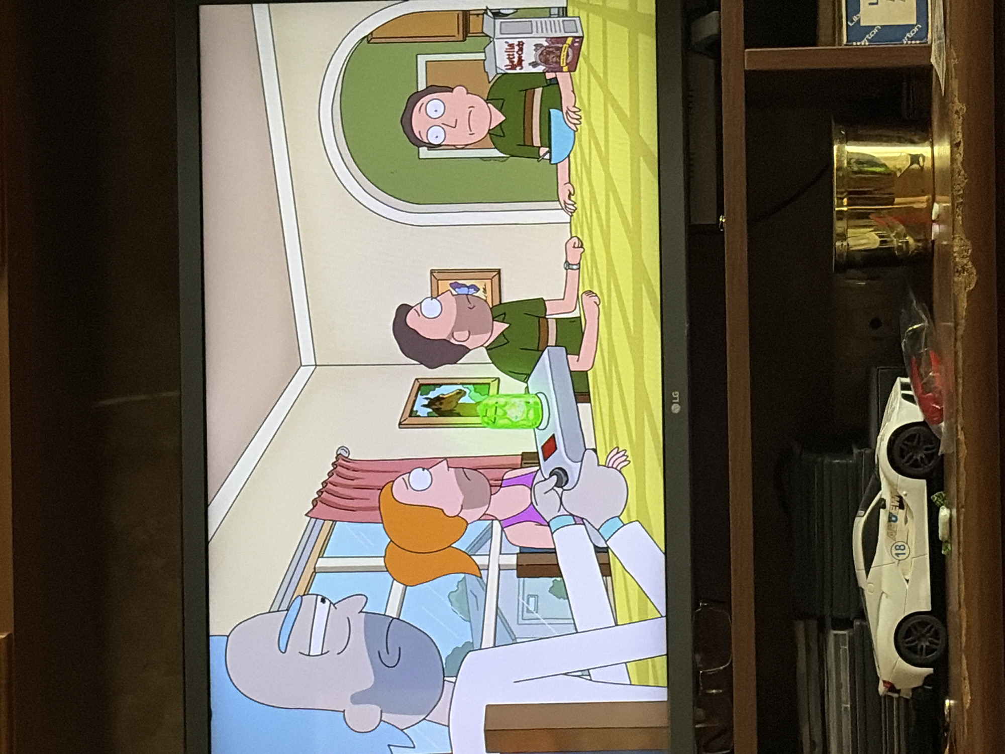 The image captures a scene from an animated television show, likely "Rick and Morty," displayed on an LG television. The scene depicts a domestic setting with four animated characters. On the left, a bald, grey-skinned character with blue irises, wearing a white lab coat, holds a grey device with a black dial. Beside him stands a female character with orange hair in a ponytail, wearing a purple tank top, and a third character, a male with brown hair, is seated at a table. A fourth male character with brown hair, identical to the third, is also seated at the table. The table is covered with a yellow tablecloth. The characters appear to be in a dining room with a window and a door visible in the background. The overall environment suggests an indoor residential setting. Below the television, a wooden shelf is visible, holding various items including a white toy car, what appear to be DVDs, a gold cylindrical container, and a box with blue text. The lighting suggests a room interior, possibly during the day.