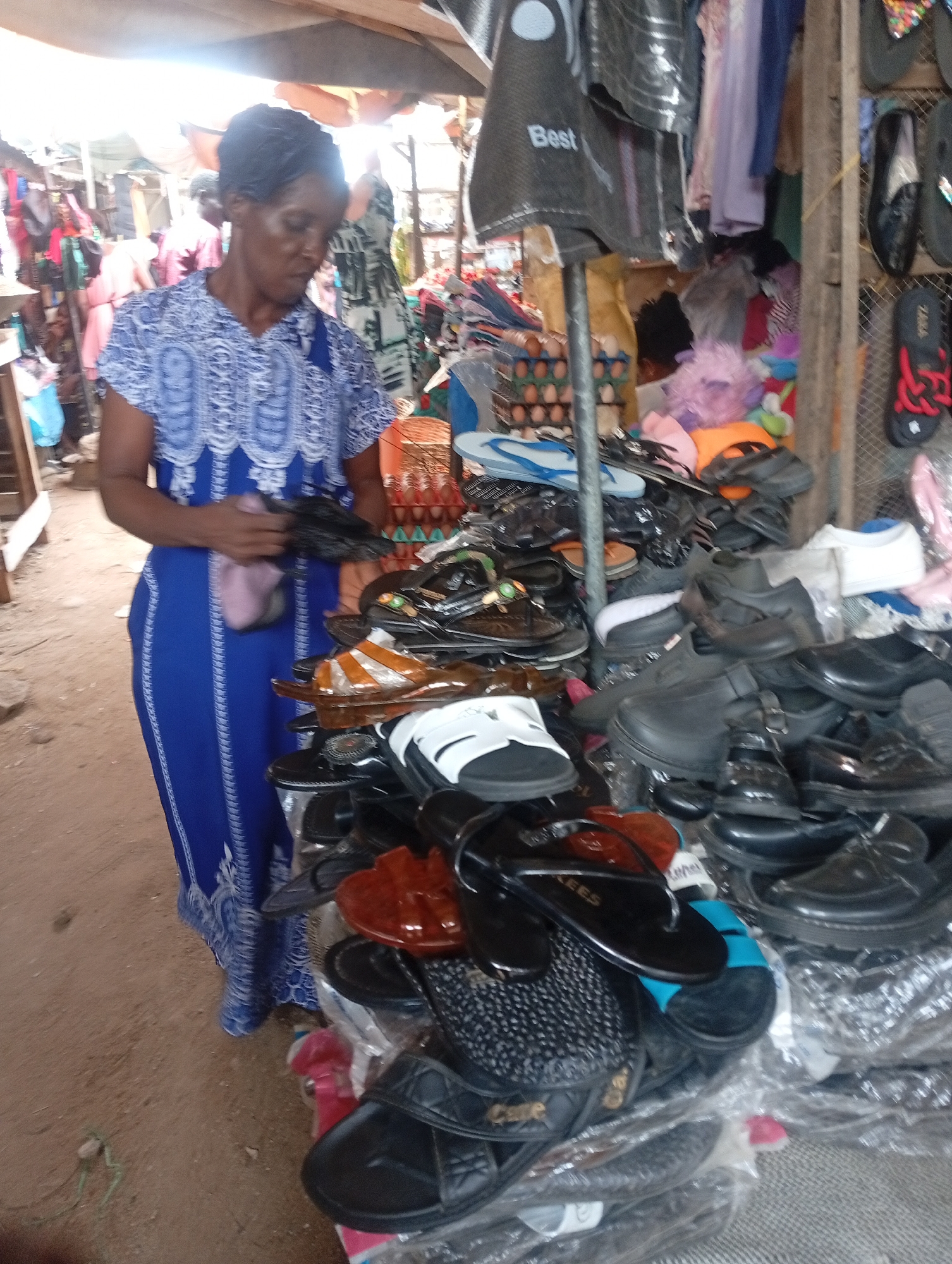 In Sabon Tasha, Nigeria, a woman in a blue patterned dress stands at a market stall densely packed with shoes and sandals. She is holding a dark-colored item, possibly footwear, and appears to be examining it. The stall is covered with a variety of footwear in different styles and colors. Behind the stall, more merchandise hangs, including clothes and accessories. Crates of eggs are visible, as well. The stall is located outdoors, beneath a makeshift roof. The ground around the stall is dirt.