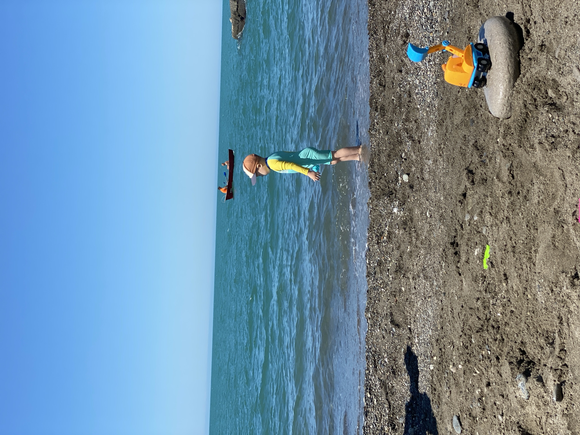 The image depicts a sunny beach scene in Sochi, Russia, characterized by a clear blue sky and turquoise-blue sea. The upper half of the frame is dominated by the unobstructed, vibrant blue sky. Below the horizon, the sea exhibits small waves, particularly noticeable near the shore where the water is shallow and transparent.
In the mid-ground, a young child stands in the shallow water, with the water level reaching just above their ankles. The child faces left, looking out towards the open sea. They are wearing a full-body swimsuit, teal/mint green on the lower half and sleeves, with a yellow torso. A beige and white baseball cap covers the child's head. The child's right hand appears to hold a small, light blue object.
Further out in the sea, to the left of the child, a small, open red boat is visible. Two individuals are seated in the boat; one in the front is wearing an orange top, possibly a life vest, and holding a paddle. To the right of the child, further out in the water, a dark, rocky outcrop protrudes from the sea surface.
The immediate foreground of the image consists of a dark grey/brown sandy beach mixed with small pebbles and shingle where it meets the water. On the dry sand in the bottom right corner, a toy orange and blue excavator with a yellow scoop is perched atop a smooth, oval-shaped grey rock, which appears wet. To the left of the image center, embedded in the sand, is a small, bright green plastic object, possibly part of another beach toy. A dark shadow, likely cast by the photographer or an unseen object, is visible on the sand in the bottom left quadrant.