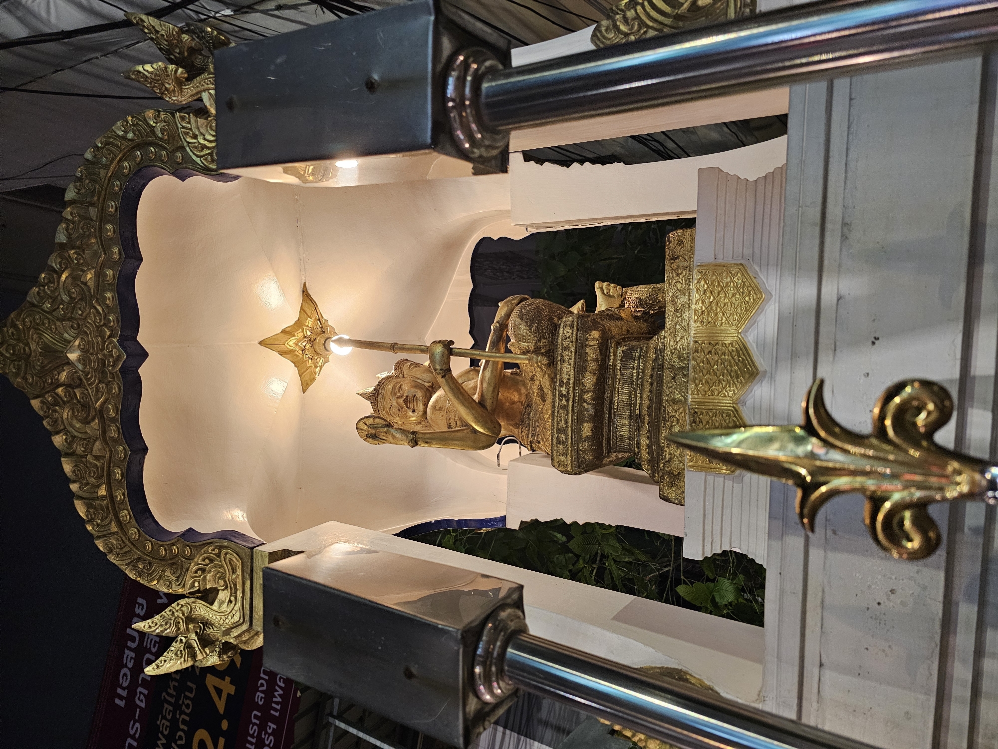 A golden statue of a multi-armed deity sits on a tiered pedestal, illuminated from above within a white shrine structure. The shrine is adorned with intricate golden carvings on its arched roof and sides, featuring dragon-like motifs. The statue is seated, holding a staff-like object and appears to have multiple faces. Two dark, rectangular pillars with metallic accents flank the shrine. In the foreground, a golden ornamental spearhead is visible, slightly out of focus. The background is dark, with hints of green foliage visible behind the shrine. A banner with Thai text and numbers is partially visible on the left side. This scene is likely located in Bangkok, Thailand, given the presence of Thai script and the architectural style often seen in religious or cultural sites in the city.
