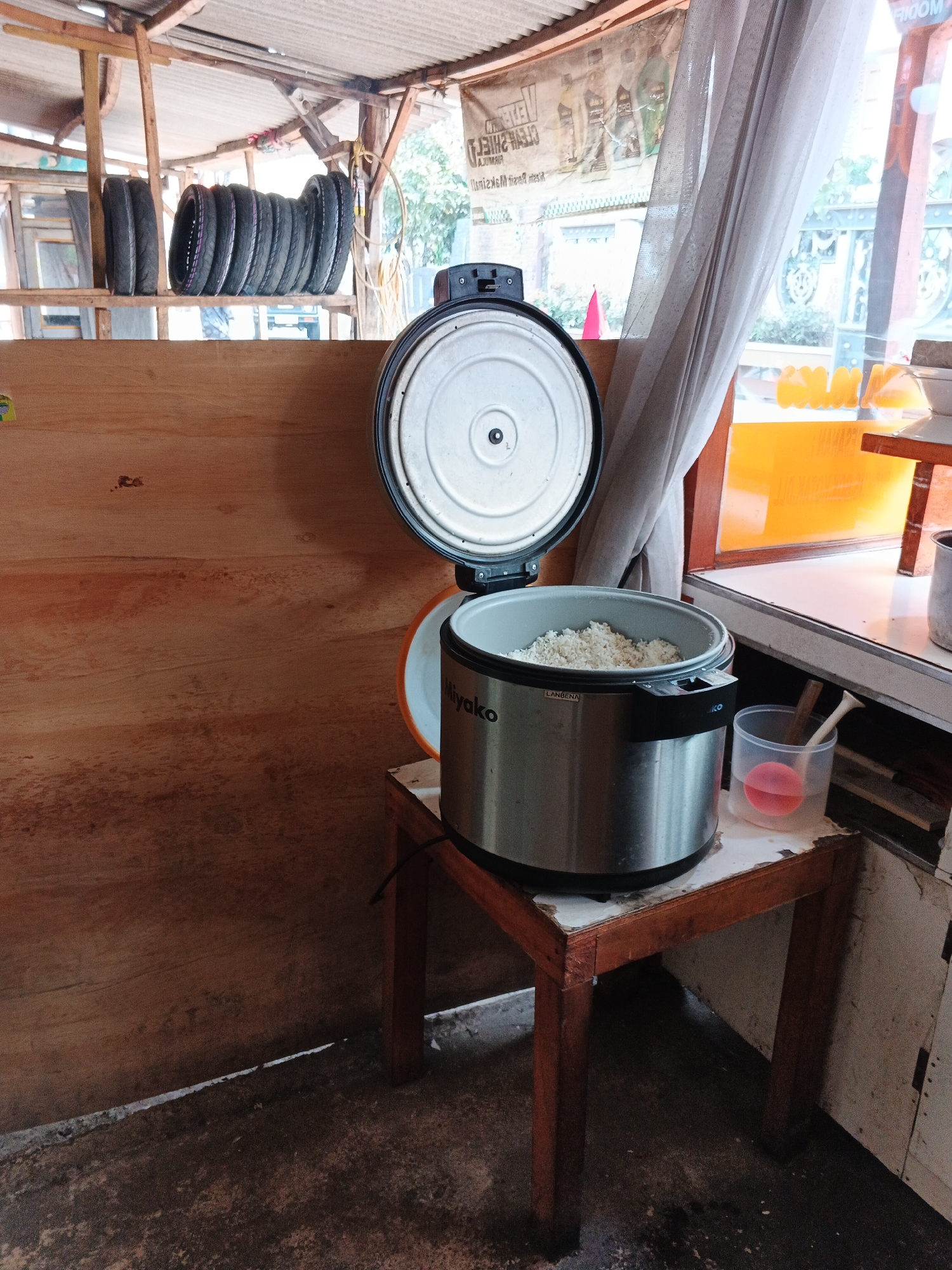 The image depicts the interior of a small, open-air establishment, likely a food stall or warung, in Gandasari, Indonesia. The dominant elements are a large, stainless steel rice cooker filled with cooked rice, placed on a small wooden stool. The rice cooker has its lid ajar, revealing the white grains within. Next to it is a clear plastic cup containing a pinkish liquid, possibly a beverage, with a wooden utensil resting inside.
The background reveals a rustic setting. To the left, stacked tires are visible, suggesting a connection to a vehicle repair service or a similar business. A wooden plank wall forms the backdrop for this area. The ceiling appears to be corrugated metal supported by wooden beams, with some translucent material allowing light to filter through. A faded banner with text in Indonesian hangs on the wall.
To the right, a window with a sheer, light-colored curtain offers a glimpse of the outside, where sunlight illuminates an orange-tinted area, possibly a serving counter or another part of the establishment. The floor is rough and appears to be concrete, with some debris scattered on it. The overall impression is one of a functional, no-frills business environment common in many Indonesian towns and villages. There are no people visible in the image.