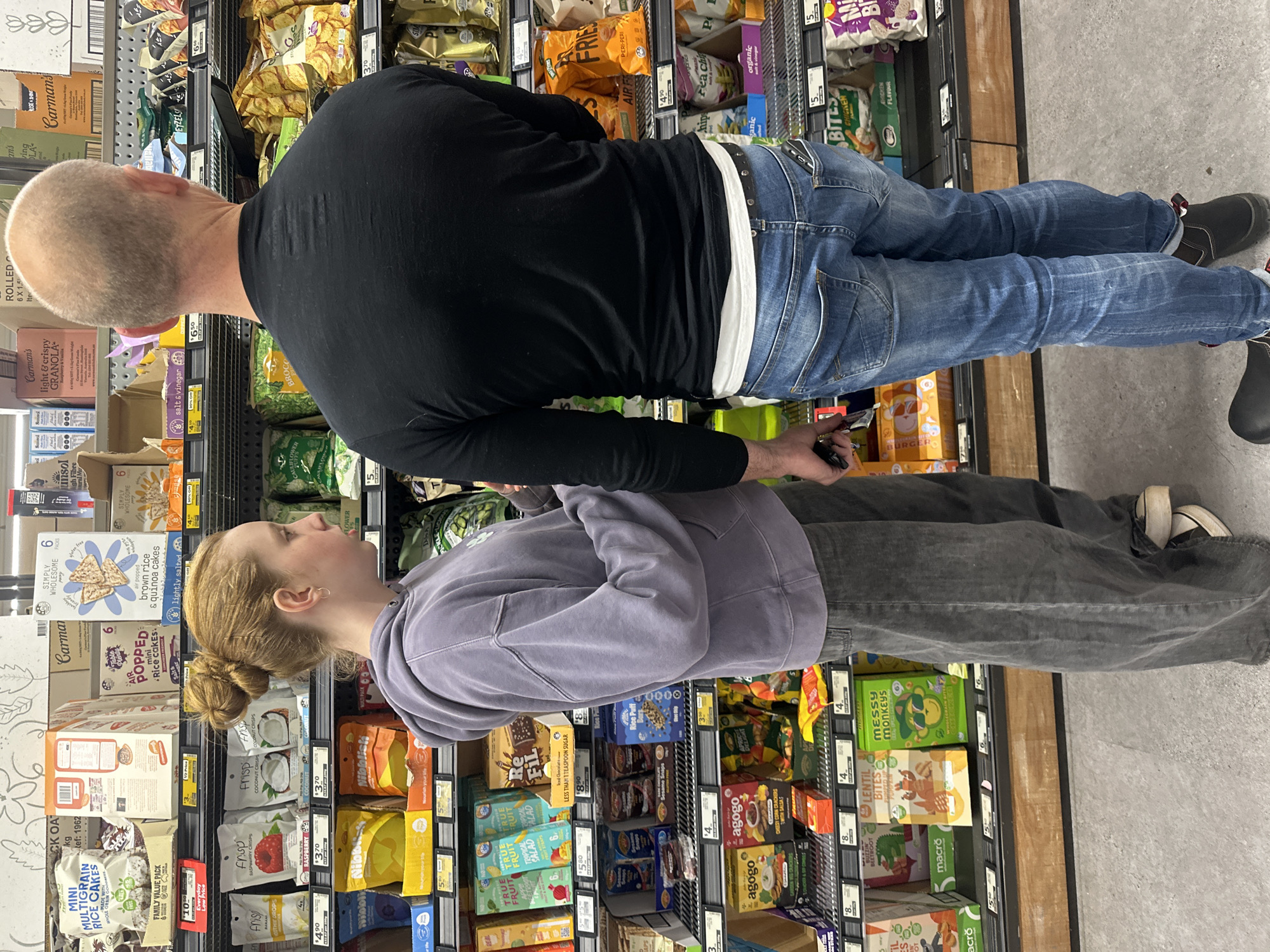 The image captures a shopping scene inside a supermarket aisle, specifically at Woolworths Toormina, Australia. A young woman with auburn hair styled in a bun, wearing a grey hooded sweatshirt and grey wide-leg jeans, stands facing away from the camera, examining a shelf of snacks. To her right, a bald man, seen from the back, is also looking at the shelves, dressed in a black long-sleeved shirt and blue jeans. Both individuals appear to be in the process of grocery shopping. The shelves are well-stocked with various packaged food items, including rice cakes, granola, chips, and other snack products, each with visible price tags. The lighting is typical of an indoor retail environment, with overhead fluorescent lights illuminating the products and aisles. The floor is a light grey concrete.