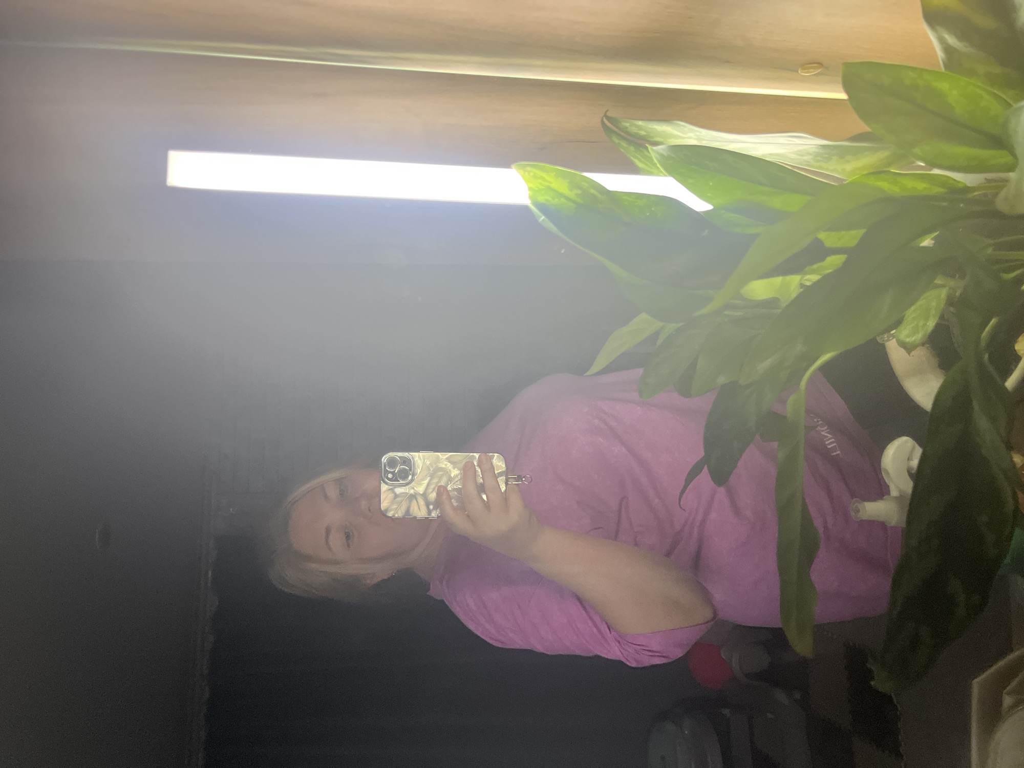 A person is taking a selfie in a mirror. The person has blond hair and is wearing a purple t-shirt. The person is holding a smartphone with a gold-colored case. A plant is visible on the right side of the frame. The background is dark. There is a fluorescent light above the person and plant. It is not possible to determine the location of the image.