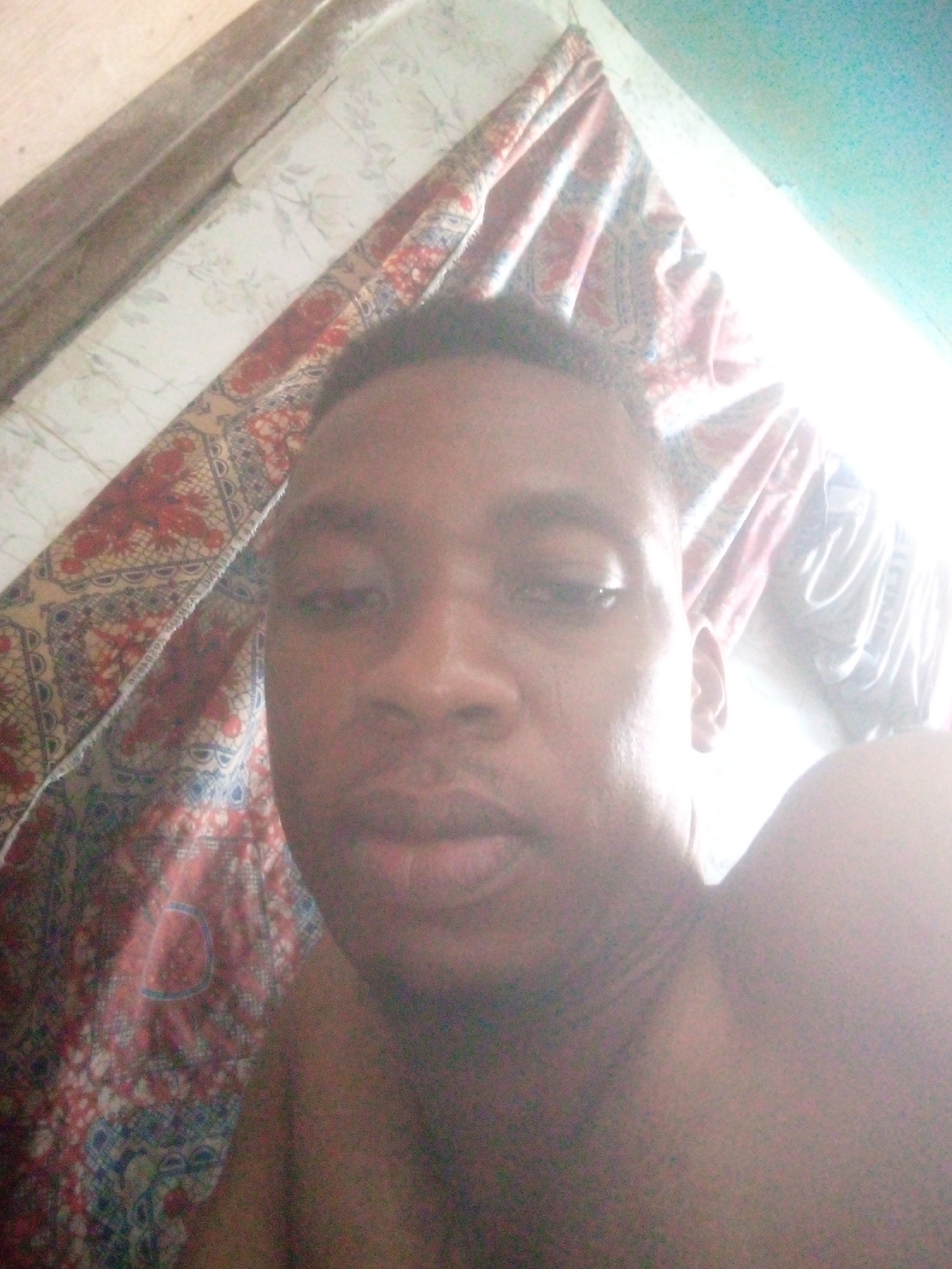 A close-up shot shows a Black man's face and upper body, presumably taken in a bedroom. The man is looking towards the camera. Behind him, a patterned curtain is visible along with a wooden frame in the upper left. The room appears to be located in Unknown, Unknown. The overall lighting in the image is bright, particularly in the top right corner, possibly indicating sunlight or an artificial light source.