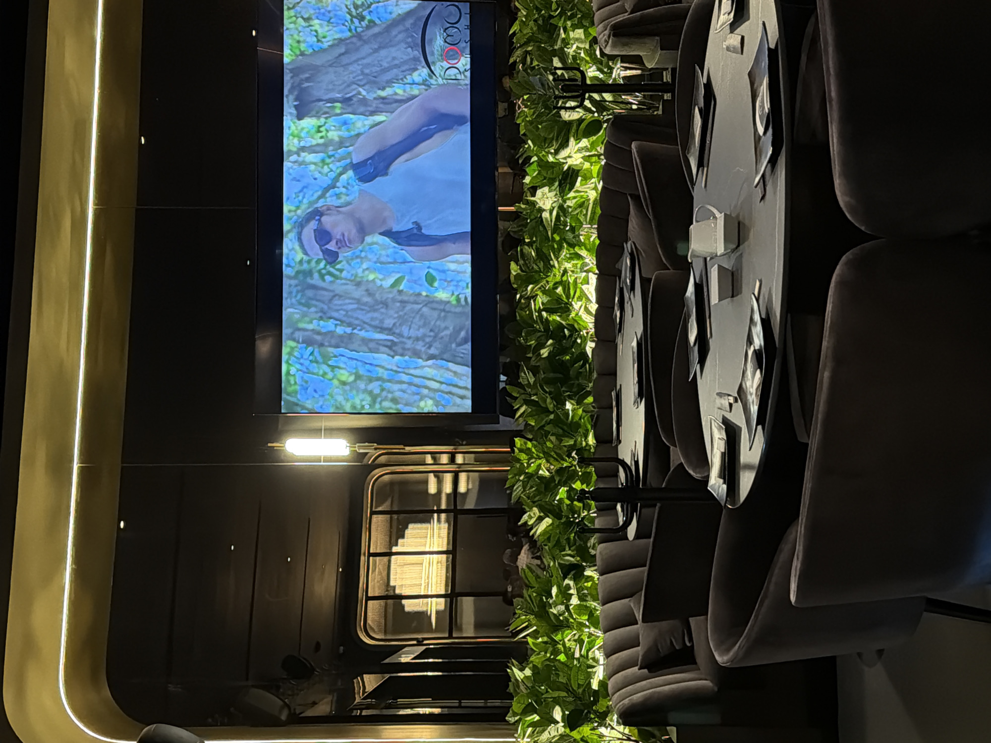 The image depicts the interior of a modern establishment, likely a restaurant, bar, or lounge, in San Bernardino Tlaxcalancingo, Mexico, characterized by a dark, sophisticated ambiance illuminated by artificial light.
The dominant feature is a large vertical screen on the left, displaying a video of a man outdoors. The man wears a light-colored top and sunglasses, set against a backdrop of blue and green foliage, suggesting a natural or hiking environment. In the top right corner of the screen's content, a small logo is visible, appearing to read "DOC" or "DOG" with a red circle enclosing the 'O'.
To the right of the screen, a long planter filled with lush green foliage acts as a natural divider, separating the wall with the screen from a seating area. This area features multiple round, dark-colored tables. Each table is surrounded by plush, dark gray upholstered chairs, arranged in a booth-like configuration along a wall. The chairs have soft, textured upholstery, possibly velvet, and appear comfortable. The tables have stylized, dark placemats or decorative elements on them.
Various lighting elements contribute to the atmosphere: a bright vertical strip light runs along a golden-toned wall section on the far left, and a horizontal bar light is positioned below the large screen. A window-like structure with a grid pattern is also visible, reflecting light sources within the room or from outside.
The setting suggests it is evening or nighttime due to the reliance on artificial lighting. No specific activity is taking place in the foreground, but the arrangement implies a space for dining, socializing, or relaxation. The overall mood is contemporary and serene. No people are actively visible in the seating area, and no specific external location cues for San Bernardino Tlaxcalancingo are present within this interior view.