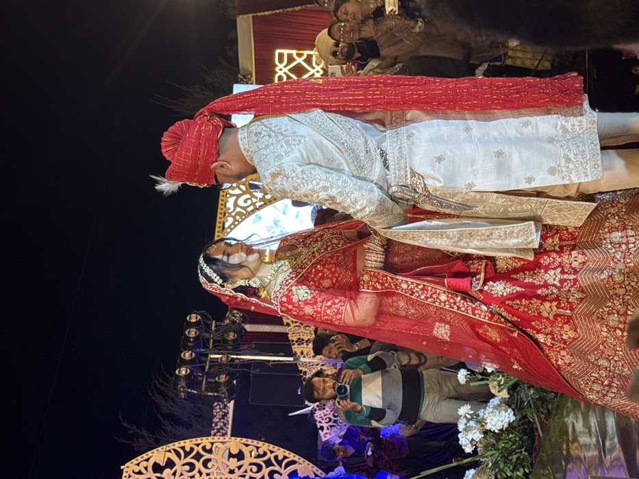 A wedding scene unfolds at night. A bride, adorned in a red and gold lehenga, stands beside the groom, who is dressed in a white sherwani and red turban. They are at the center, with photographers capturing the event. The ornate wedding stage is visible in the background.