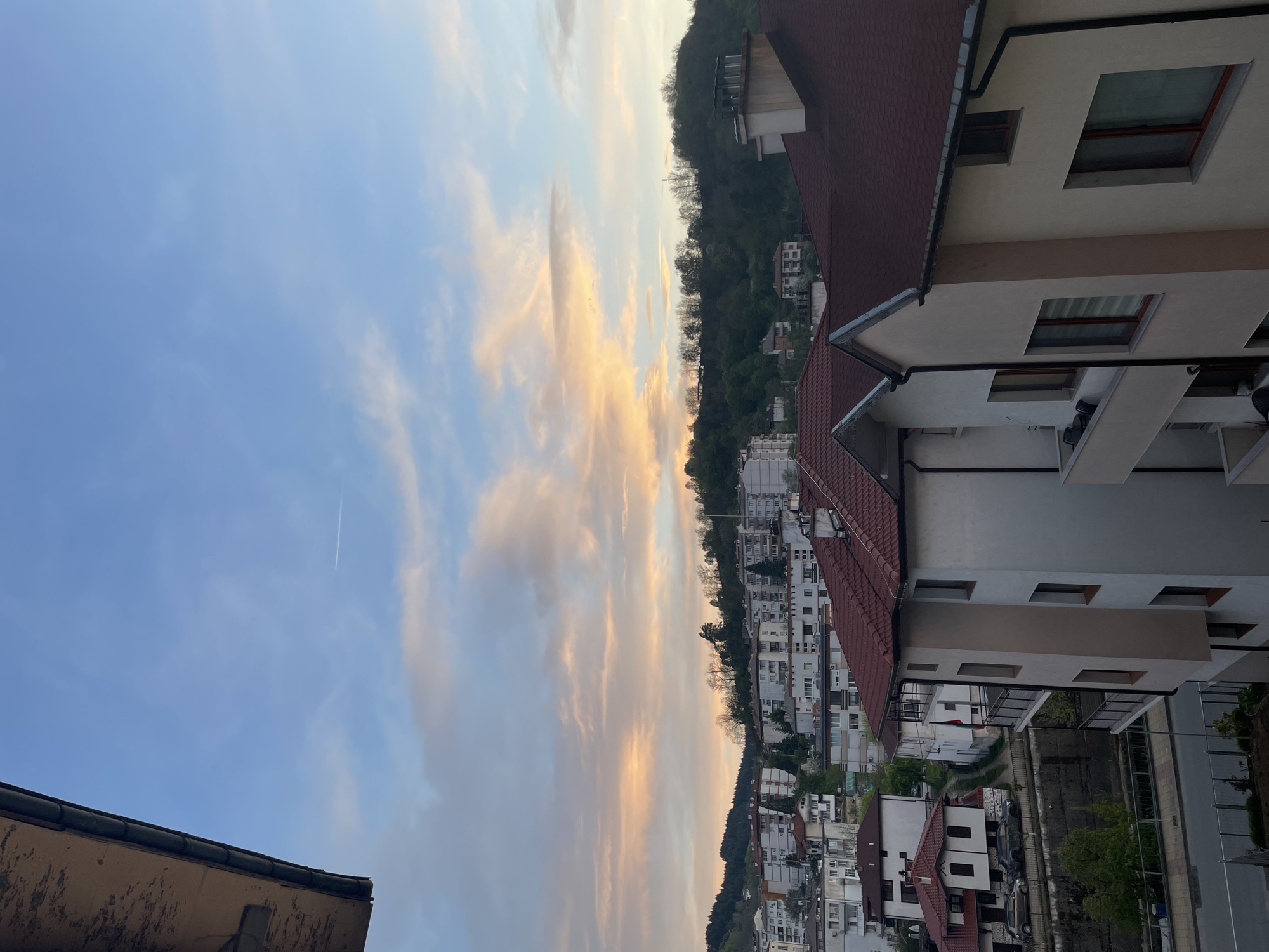 The image shows a cityscape in Zlatograd, Bulgaria, viewed from an elevated position. The sky is partially cloudy with hints of yellow and orange. Buildings with red-tiled roofs and light-colored facades dominate the right side of the frame, descending into a valley. On the opposite side of the valley, a tree-covered hill rises, dotted with houses and buildings. A contrail from an aircraft is visible in the upper left portion of the sky.
