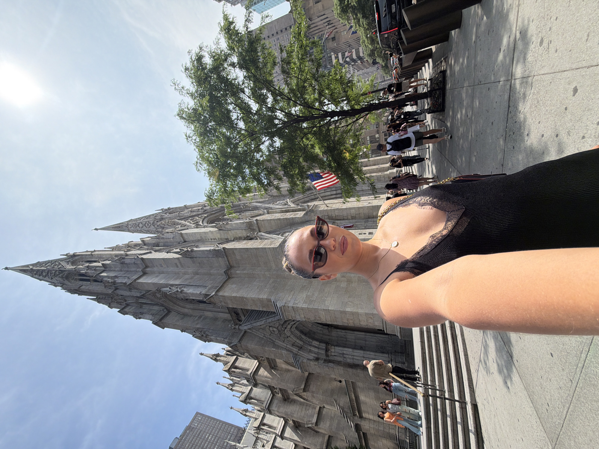 A woman takes a selfie in front of St. Patrick's Cathedral, a landmark in Midtown Manhattan, New York City. The cathedral is made of granite and features Gothic Revival architecture with soaring spires and intricate carvings. The woman is wearing sunglasses and a black dress. Other people are visible walking on the sidewalk and climbing the steps of the cathedral. A tree with green leaves stands to the right of the cathedral, and an American flag is visible on a pole. The sky is blue with some clouds. The scene appears to be a sunny day in the city.