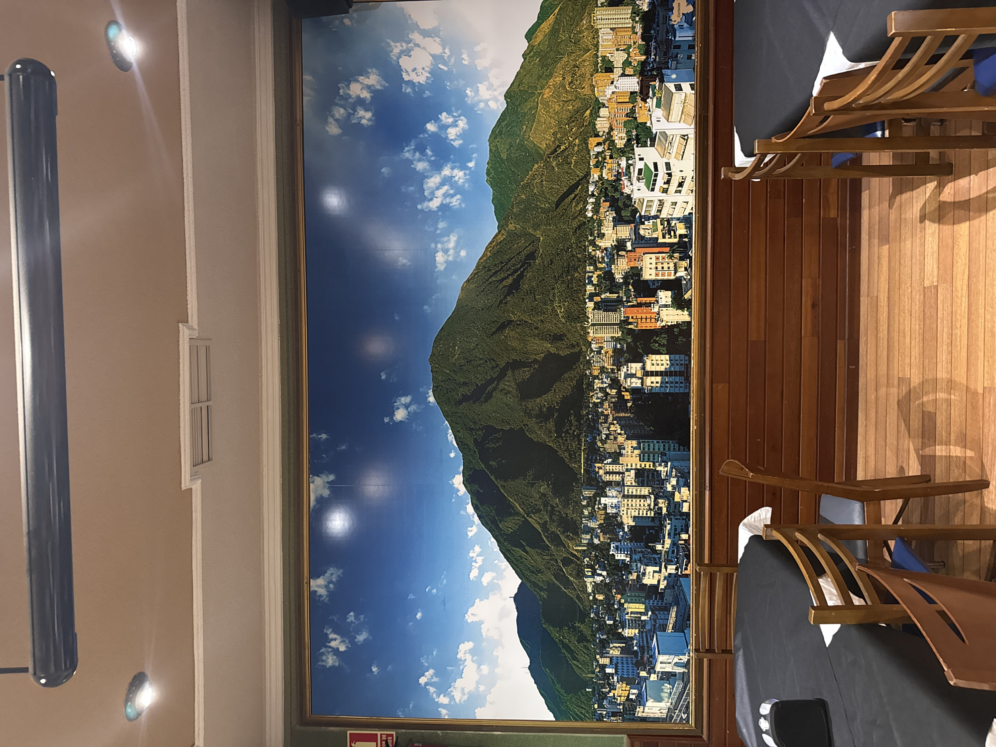 The image depicts an indoor dining area with a large mural of a city and mountains covering the back wall. The mural shows a dense urban landscape with numerous buildings of varying heights and colors, set against the backdrop of lush green mountains under a bright blue sky with scattered white clouds. The mural is framed by a golden border. Below the mural, the wall is paneled with horizontal wooden planks. In the foreground, two round tables are set with black tablecloths and white napkins. Several wooden chairs with blue seats are placed around the tables. The floor is made of light-colored wooden planks. In the upper part of the image, a dark, cylindrical fixture, possibly a projector or light, is mounted on the ceiling, along with two recessed circular lights. A red fire extinguisher is partially visible on the left edge of the mural. The overall setting suggests a restaurant or dining hall. The location of Torrejón de Ardoz, Spain, is not discernible from the visual information present in the image.