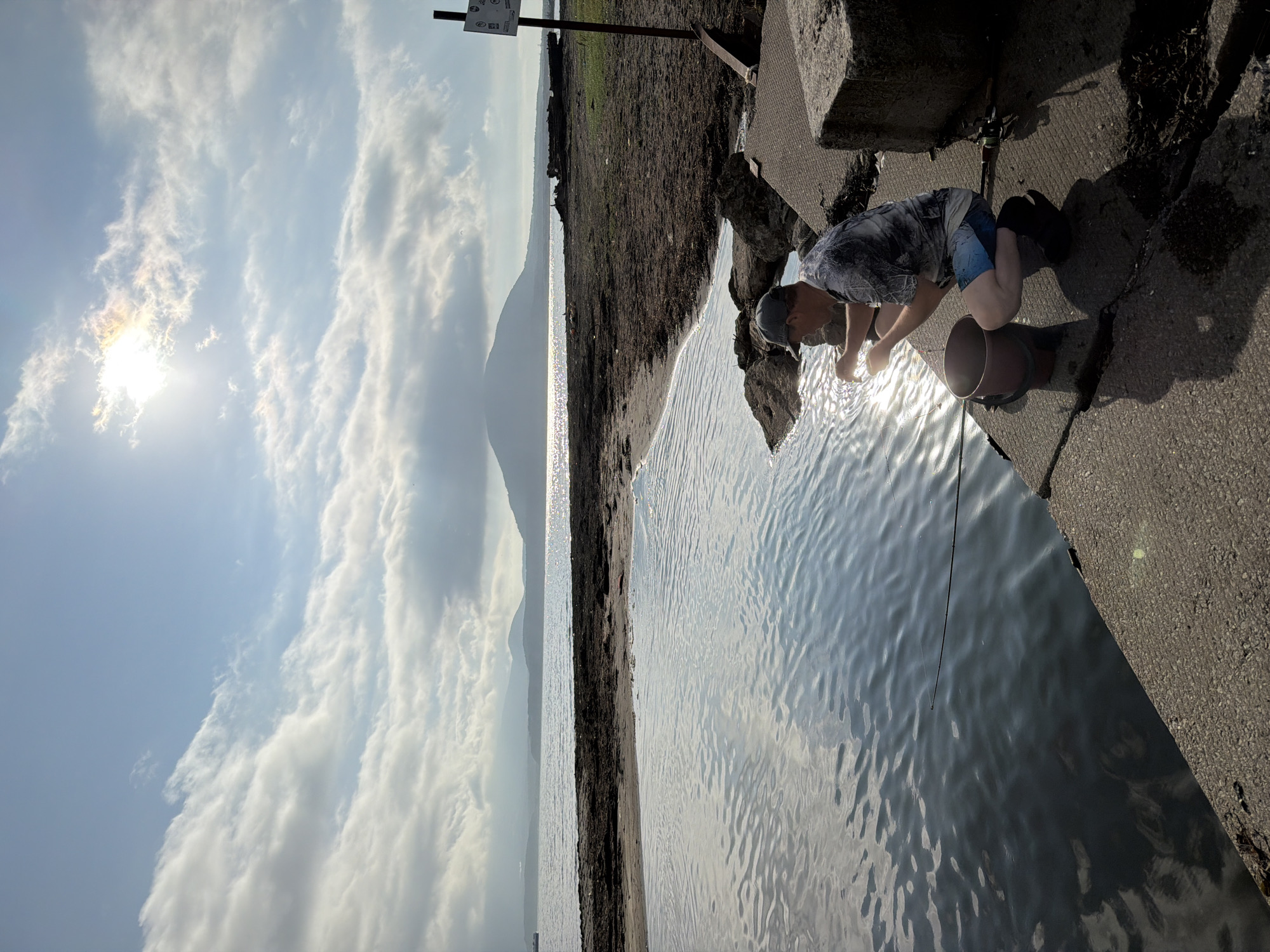 An individual is fishing on a concrete pier overlooking a body of water. The person, wearing a grey baseball cap, a patterned t-shirt, and blue patterned shorts, is crouched down. In front of them is a bucket and a fishing rod. The sun is shining brightly, casting reflections on the water and creating shadows on the pier. In the background, there is a sandy shore and a distant landmass with a mountain. The sky is partly cloudy.