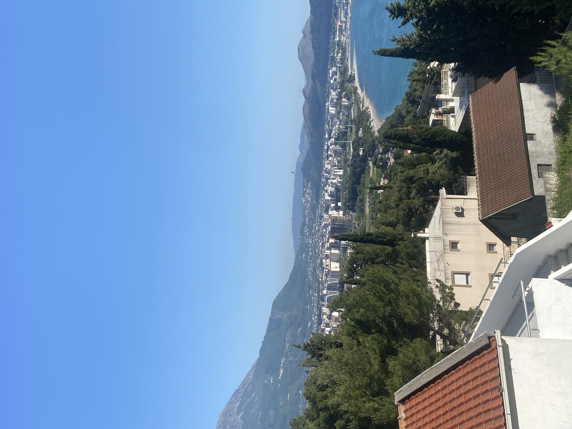 The image shows a cityscape of Šušanj, Montenegro, viewed from an elevated perspective. In the foreground are the rooftops of buildings, with a mixture of brown tile and white painted surfaces. Verdant trees fill the space between the buildings. The cityscape consists of dense buildings stretching along the coastline. A narrow beach runs along the shore where the city meets the Adriatic Sea. Mountains rise in the background, partially obscured by atmospheric haze. The sky is clear and bright blue.