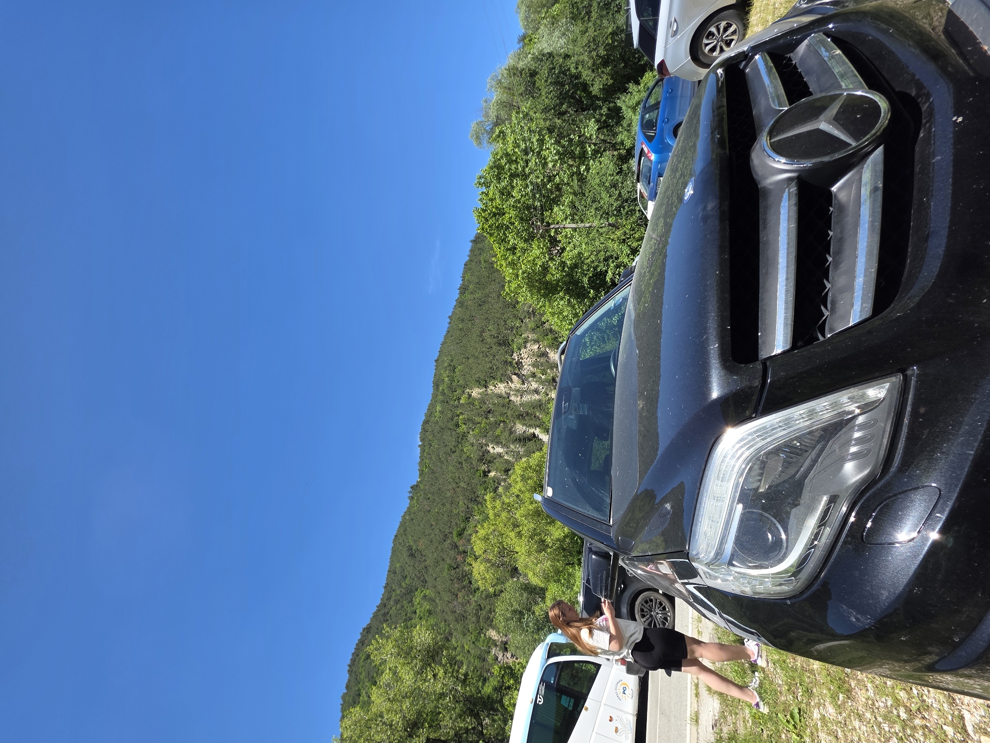 A black Mercedes-Benz SUV dominates the foreground, parked on the roadside in Bachkovo, Bulgaria. A woman stands next to the vehicle. Behind the car and woman, a white van and a wooded hill slope. The sky is clear and blue.
