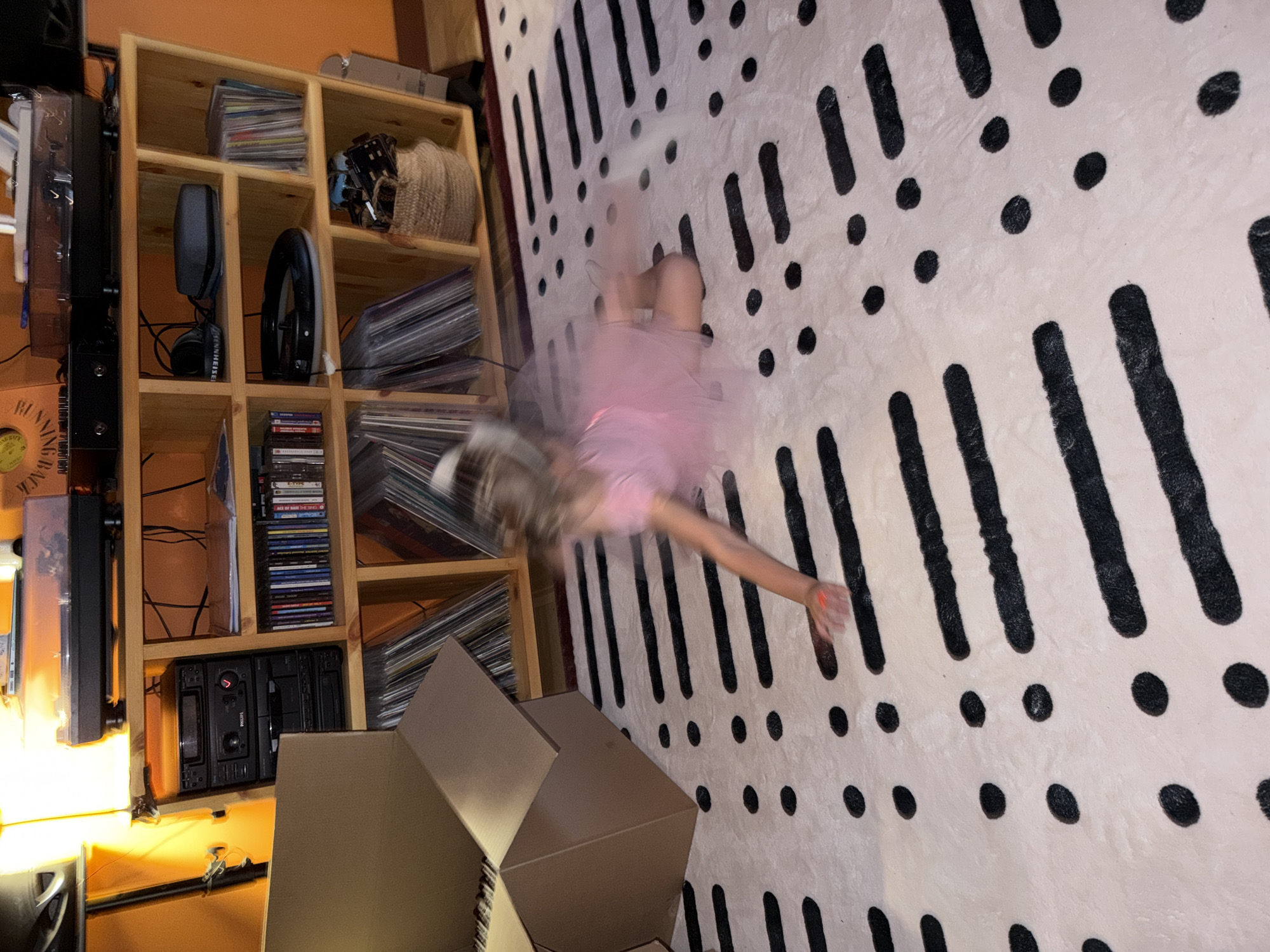 A young child, blurred with motion, is captured mid-fall on a cream-colored rug with a black abstract pattern of lines and dots. The child is wearing a pink tutu and appears to be tumbling or dancing. The scene is an indoor setting, likely a living room or playroom, given the presence of a wooden shelving unit filled with various items.
On the shelves, there are stacks of CDs, what appear to be vinyl records, a pair of headphones, a circular object resembling a speaker, and a woven basket. To the left of the shelves, a tall black electronic device with glowing orange lights is visible. In the foreground, partially obscuring the shelves and the rug, are two large cardboard boxes. One is fully assembled and open, while the other is partially collapsed.
The lighting suggests it might be evening or an indoor space with artificial light, as indicated by the glow from the electronics and the overall illumination. The child's blurred movement evokes a sense of playfulness and energy, perhaps a spontaneous dance or a moment of exuberant activity. The setting appears somewhat cluttered, suggesting a lived-in space. No specific text is clearly legible on any of the objects, though some CD cases and record sleeves might have writing that is too small or blurred to read. The overall impression is one of a candid moment of childhood play within a home environment.