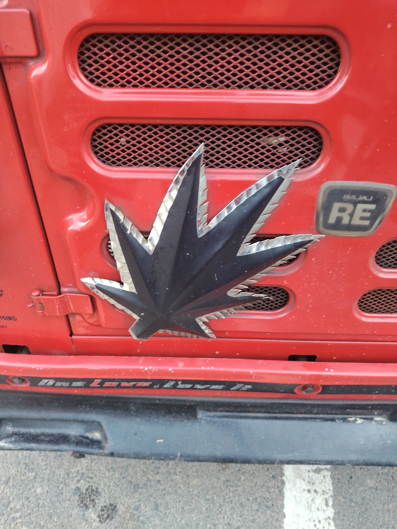 This is a close-up, eye-level, outdoor photograph of the front of a red Bajaj RE vehicle. The vehicle has a silver and black cannabis leaf emblem affixed to its front. To the right of the emblem is a badge that reads "BAJAJ RE". The vehicle is parked on a textured, grey surface with white markings, possibly a road or parking lot. The image is well-lit and in focus.