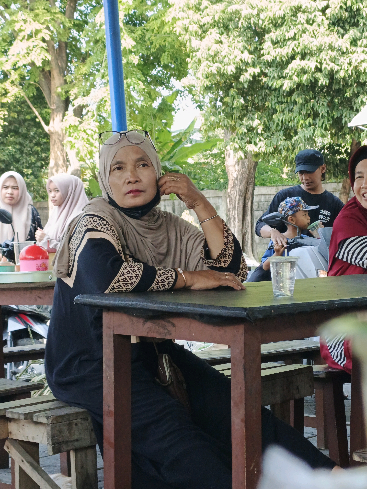 In the city of Kludan, Indonesia, a group of individuals is seated at outdoor tables under a canopy. A woman in the foreground, wearing a beige hijab and a black and patterned outfit, rests her chin on her hand, her glasses perched on her forehead. To her left, two younger women in hijabs are visible. To the right of the foreground woman, a man wearing a black cap and shirt, and a young boy in a colorful cap are seated. A clear plastic cup with a beverage sits on the table in front of the foreground woman. The background consists of lush green trees and a concrete wall. The tables and benches appear to be constructed from wood and metal, suggesting a casual, possibly food stall or eatery setting.