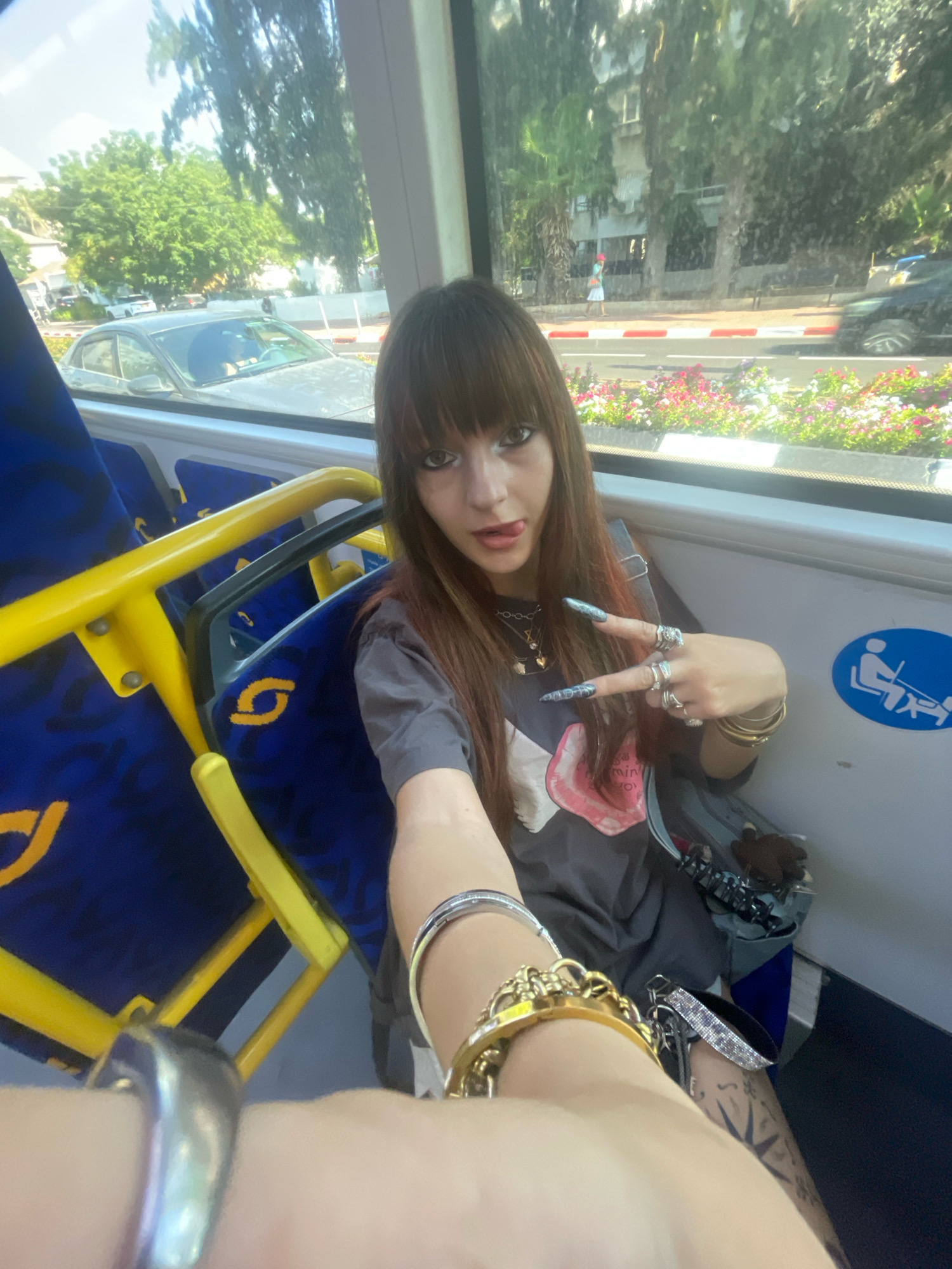 The image shows a young woman with dark hair and bangs sitting on a bus in Raanana, Israel. She is wearing a grey t-shirt with a graphic design and multiple rings on her fingers, including a silver band on her left index finger and a large gold bracelet on her right wrist. She is holding her right hand up, forming a peace sign, and sticking her tongue out. The bus seats are blue with a yellow handrail. Through the bus window, trees, buildings, cars, and a flower bed are visible, suggesting an urban environment. A blue sign with a white icon depicting a person with a cane is affixed to the wall of the bus.