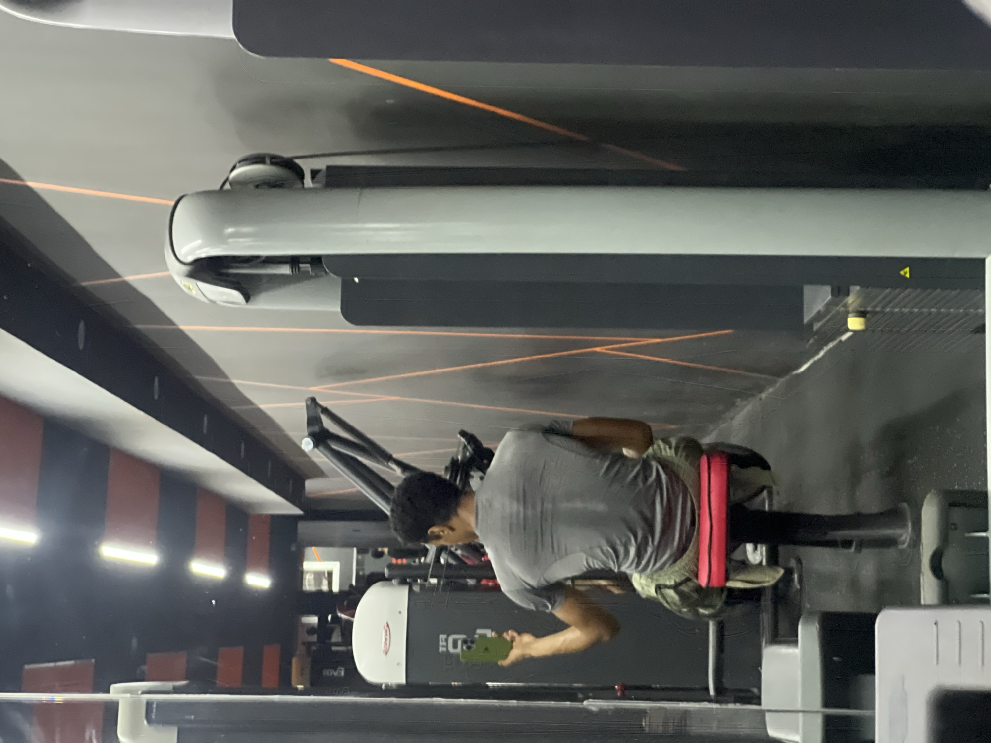 The image, rotated 90 degrees clockwise, depicts an indoor gym scene in M'Diq, Morocco. The primary subject is a person, likely male, seen from a rear-side perspective, seated on a piece of gym equipment, possibly a rowing machine. He has dark hair and is wearing a short-sleeved grey t-shirt, green or camouflage-patterned shorts, and a distinctive red band around his waist.
The person is actively looking at and holding a bright green smartphone in his right hand, suggesting he is taking a break from his workout, checking messages, or using a fitness application.
The gym environment features various exercise machines. Parts of a large treadmill with grey and black components are visible in the foreground and to the person's right. Other gym equipment, including a machine with a white panel on which partially visible black text resembling "EOD" or "EOV3" is present, can be seen in the background. The gym's interior design includes dark walls or panels with thin, striking orange lines, possibly decorative lighting or reflective elements. Bright horizontal strip lights are reflected on a dark, glossy surface, contributing to the artificial lighting of the space. A faint yellow triangular warning symbol is also visible on a dark surface in the background. The scene conveys a moment of pause or casual activity within a workout setting.
