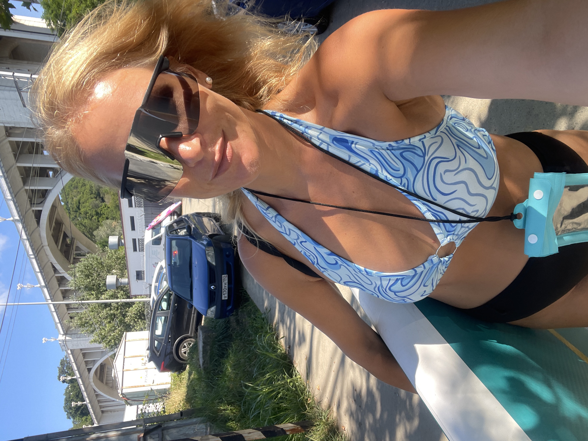 In Sochi, Russia, a fair-skinned woman with blonde hair, wearing oversized reflective sunglasses, a blue and white patterned bikini top, and black bikini bottoms, poses for a selfie while holding a paddleboard. She is wearing a translucent blue waterproof pouch on a lanyard around her neck. The background features a concrete bridge with arched supports spanning over a wooded area. Several cars are parked near a building with a white shipping container. The scene is illuminated by bright sunlight, casting shadows on the ground.